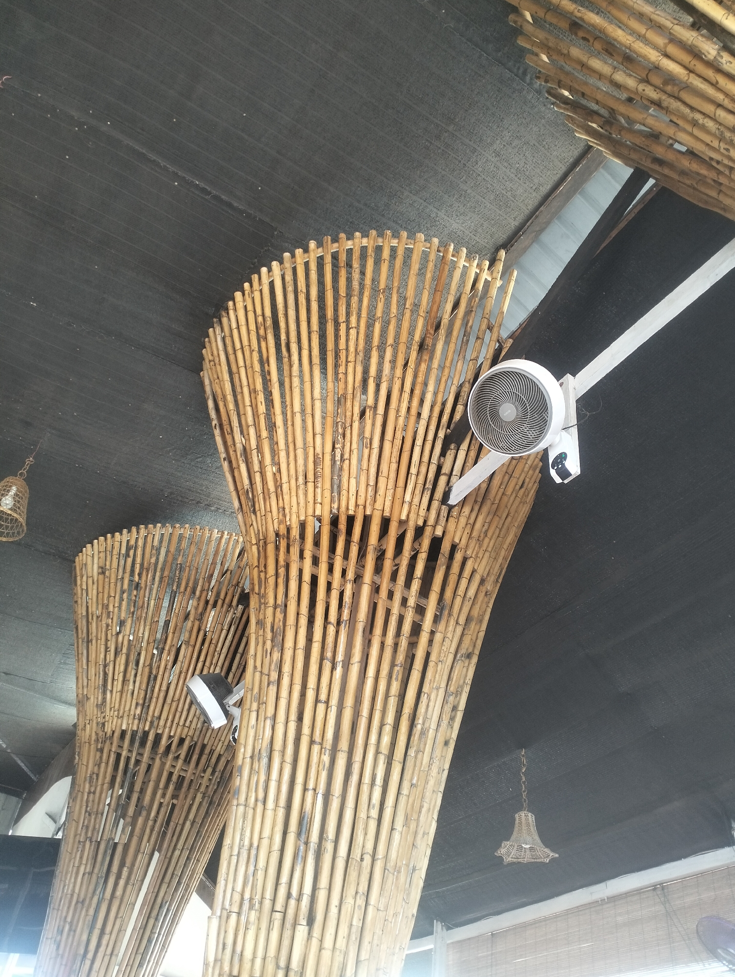 In Uteungeulinggang, Indonesia, the image captures a ceiling decorated with bamboo structures. Two large, conical bamboo installations are prominent, with their vertical poles creating a textured, organic form. A white ceiling fan is mounted on a white bracket attached to a white pole extending from the ceiling, positioned above the bamboo. A second, smaller fan is mounted on a white bracket attached to the bamboo structure itself. A small, woven lampshade hangs from a chain on the ceiling. The ceiling is covered with a dark, textured fabric. The overall scene suggests an outdoor or semi-outdoor space, possibly a restaurant or cafe, with a natural, rustic ambiance.