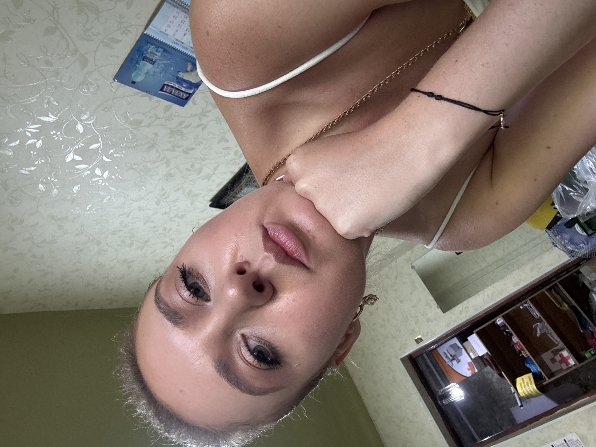 A close-up, slightly upward-angled selfie features a young Caucasian woman with blonde hair pulled back and light makeup. She is wearing a white strap top and gold jewelry, including a pendant necklace and a hoop earring with a stone. Her left hand rests on her chin, and her right arm is bent, with a black string bracelet visible on her wrist. The background shows a shelf with various items, including a red cross symbol, a fan, and some bottles, along with a patterned wallpaper. A calendar with an image of a polar bear is visible in the lower right corner. The lighting suggests an indoor setting.