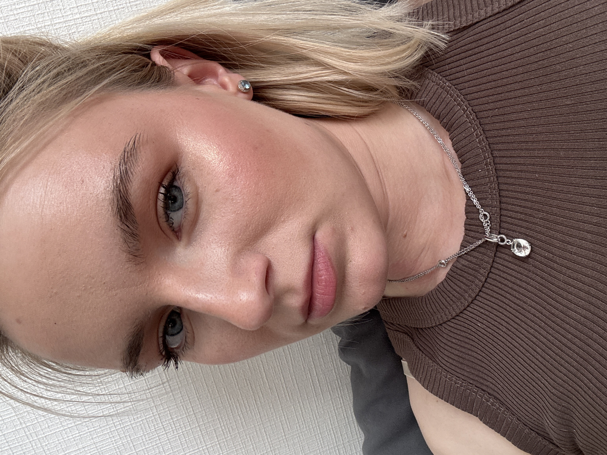 A close-up portrait of a woman with blonde hair, blue eyes, and fair skin is presented at an angled view. She is wearing a brown ribbed top and a silver necklace with a clear teardrop-shaped pendant. A silver stud earring is visible in her left ear. The background includes a white textured surface and a dark gray fabric. The location is not identifiable from the image.