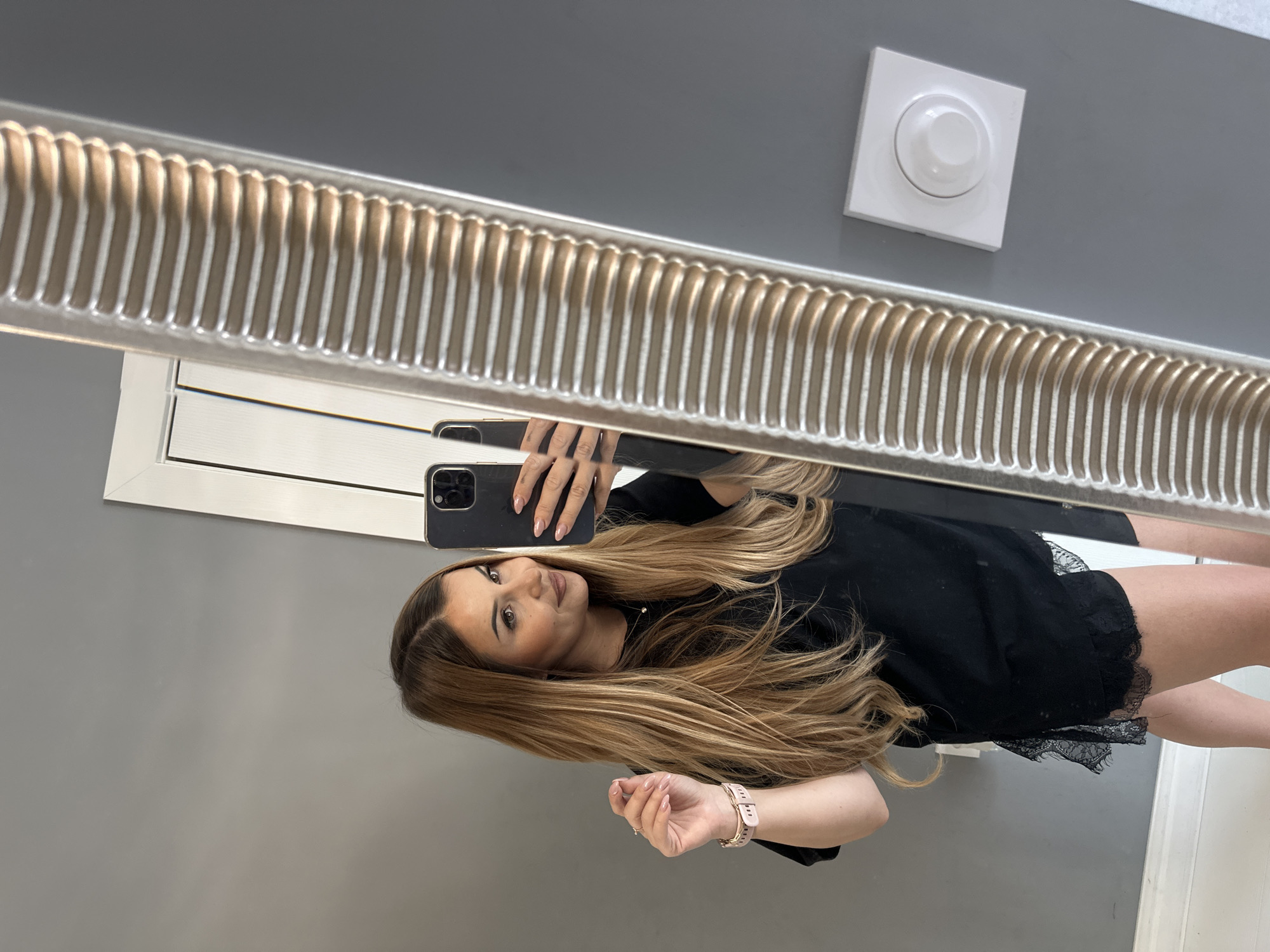 In the city of Asenovgrad, Bulgaria, a female individual takes a mirror selfie indoors. The subject, a woman with long, multi-toned hair featuring darker roots and lighter caramel-blonde ends, is positioned facing a large mirror that dominates the right side of the frame. She looks towards her reflection with a slight smile. Her left hand is slightly raised, displaying a light pink smartwatch with a ribbed strap on her wrist, and her fingernails are painted a pale pink/nude color. In her right hand, she holds a black smartphone with a visible multi-lens camera array, pointing it towards the mirror to capture the image.
The woman is wearing a loose-fitting black top, which appears to have a lace-trimmed hem, suggesting it could be part of a sleepwear set or a casual garment with decorative elements. A small, delicate necklace with a pendant is visible against her top. The background behind her reflection consists primarily of a smooth, medium-gray wall. To the upper left of her reflection, a vertical section of a white door frame is visible. At the bottom of the wall, a white skirting board is present. To the right of the reflected mirror frame, a white square electrical fixture with a central circular, translucent white knob, resembling a dimmer switch or thermostat, is mounted on the gray wall. The mirror itself features a thick, ornate frame with a repetitive, ribbed, metallic-colored texture.
