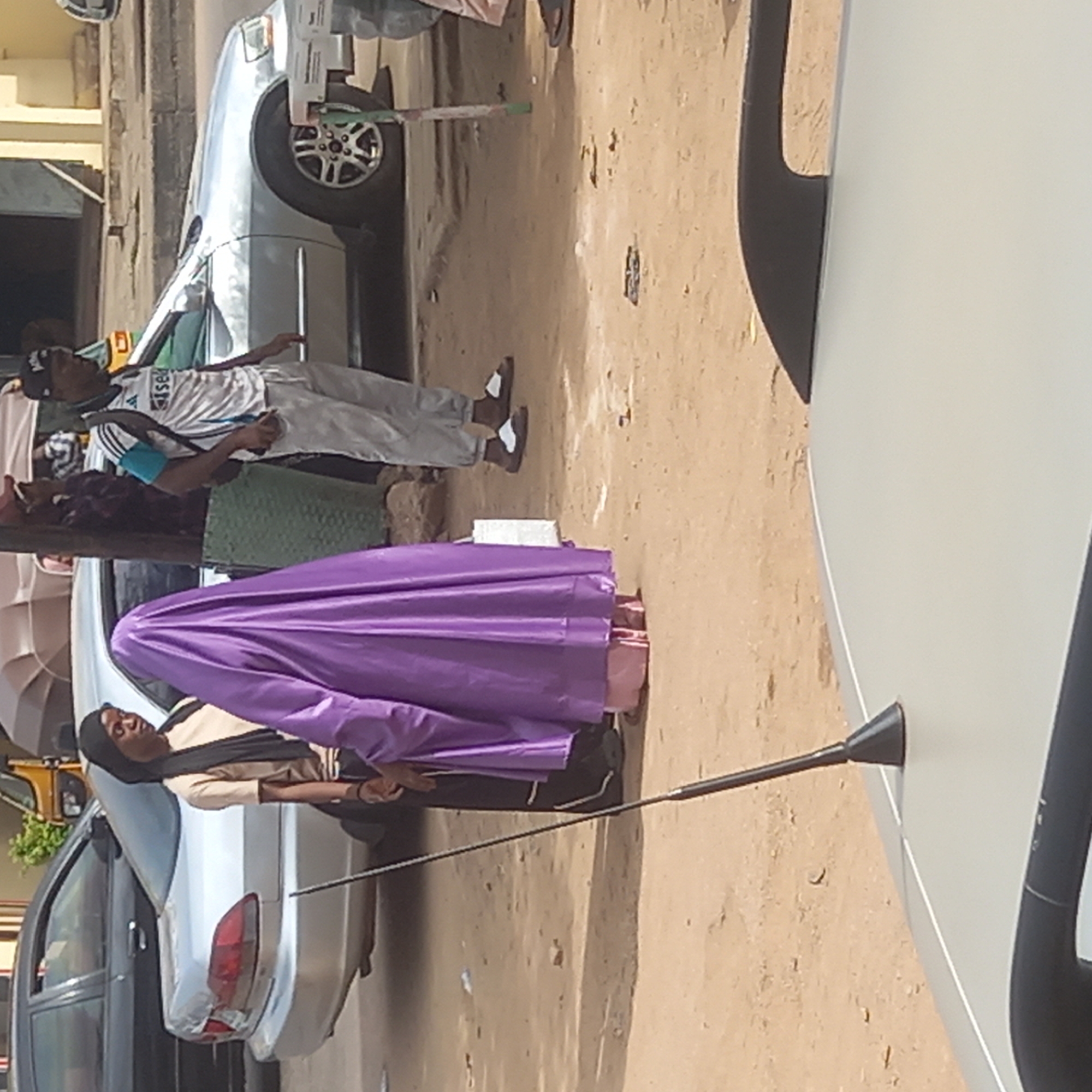 The image shows a street scene in what appears to be the city of Dadi Riba, Nigeria. A woman in a long purple robe stands between two parked cars. A man wearing a sports jersey, light-colored pants, and sandals stands nearby, along with another person wearing a dress, who is closer to the woman in purple. A man with a dark head covering stands with his back to the first car. There is a white box or package located on the ground near the woman. The ground appears to be dusty or sandy.
