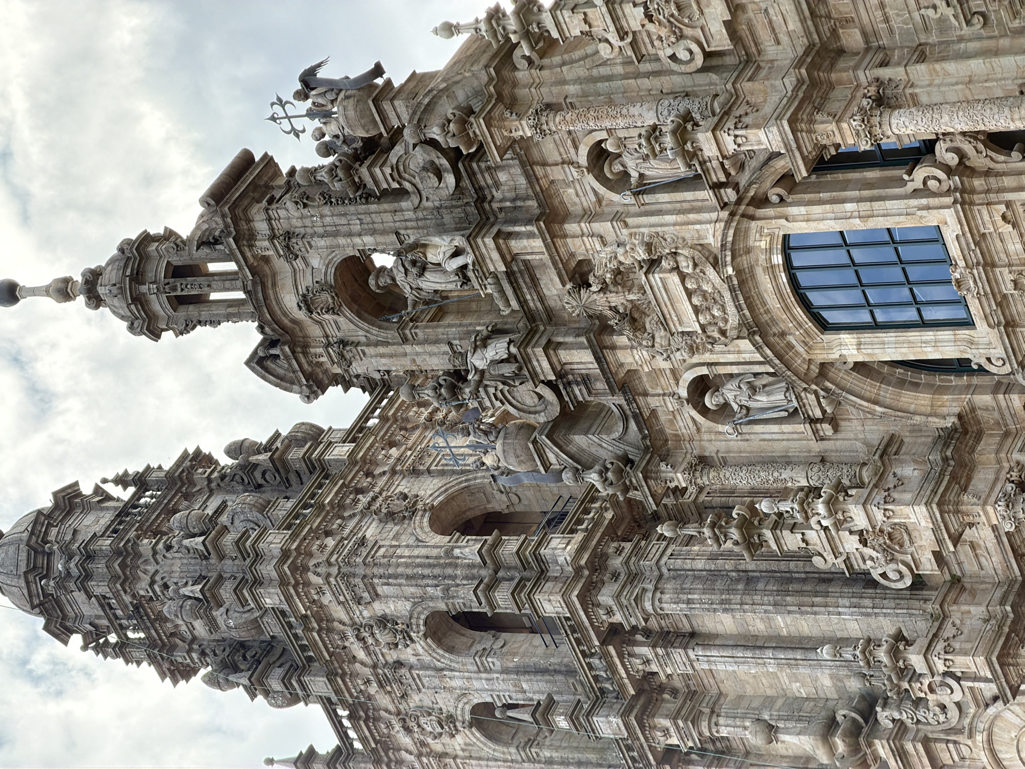 The image is a close-up, low-angle shot of the Baroque facade of the Cathedral of Santiago de Compostela in Santiago de Compostela, Spain. The detailed stonework of the cathedral's towers and ornamentation is prominent, with multiple niches housing statues of figures in robes and holding staffs or crosses. Sculptural elements, including cherubs and intricate decorative motifs, are visible. A large, multi-paned window with dark framing is centrally located in the lower portion of the image, reflecting the overcast sky. The architectural style is characterized by its grandeur, elaborate carvings, and towering structures, indicative of Baroque religious architecture. The sky above is cloudy and bright.