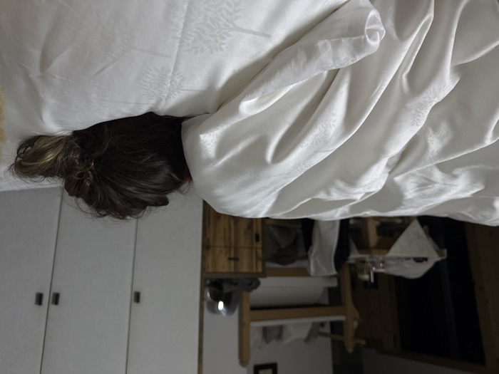 The image depicts an individual lying in a bed within a dimly lit indoor environment, identified as a bedroom. The person is positioned face down or on their side, with only the back of their head and upper body visible, covered by a white duvet and resting on a white pillow. The bedding features a subtle, embossed pattern resembling foliage or trees. The individual has long, dark brown hair styled into a messy bun or ponytail at the back of the head.
In the background to the left, a dark wooden table or bench is present, topped with a light-colored, patterned cloth and several clear glassware items. Behind this, part of a wooden bunk bed frame, with the upper bunk made up, is visible. To the right in the background, a wall features three white wardrobe doors with dark vertical handles. Adjacent to the wardrobes, a small wooden cabinet or dresser holds a reflective, curved metallic object. The specific location within Kastelruth - Castelrotto, Italy, is not visually identifiable from the image content.