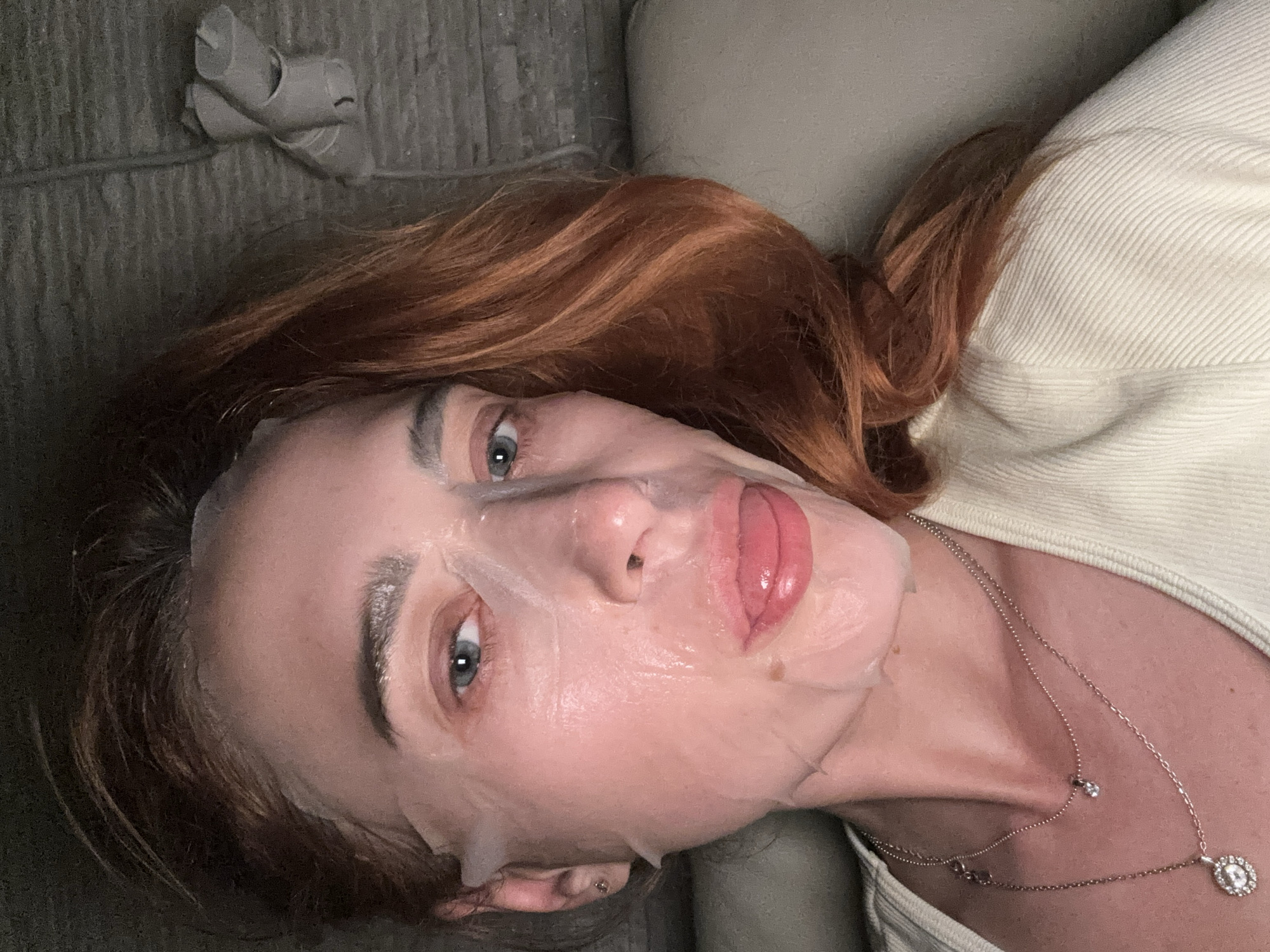 A woman lies on a grey upholstered couch or daybed with part of a beige-colored cushion visible in the upper right corner. She is wearing a sheet mask on her face and a white top, and her red hair is fanned out around her head. A silver necklace with pendant and a smaller drop is visible on her chest. Beside her, on the couch, are three empty toilet paper rolls. The image does not provide enough information to determine if the picture was taken in the city of Budva, Montenegro.