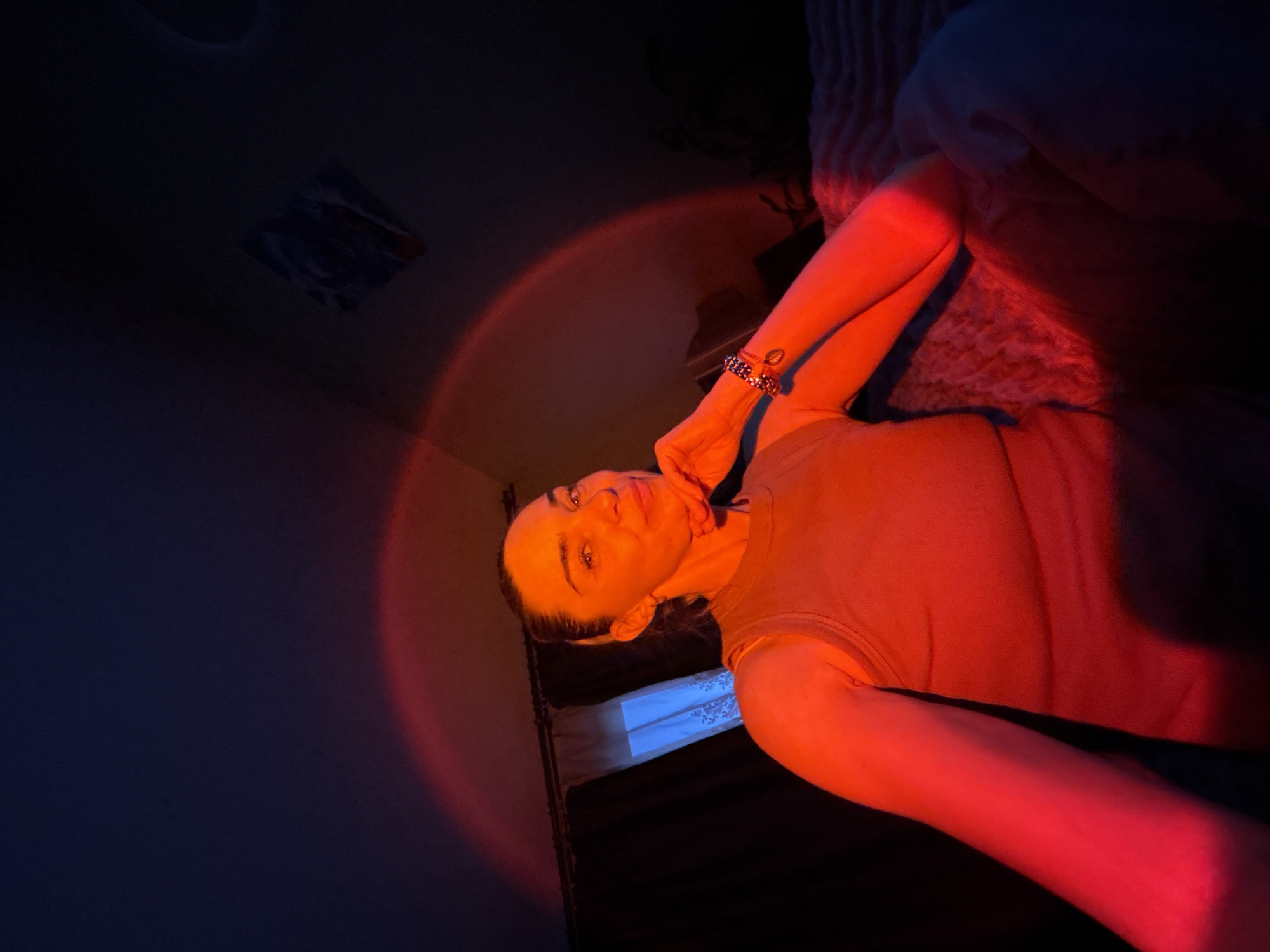 A medium shot captures a woman indoors, bathed in the warm glow of red and orange light, possibly from a sunset lamp. She has her right hand resting on her chin, and her left arm is bent, with her hand reaching towards the camera. She wears a sleeveless, tan top and multiple bracelets on her left wrist. Her hair is pulled back into a ponytail. The background is dimly lit, showing a dark curtain to the left and a window with a blue light filtering through. A textured surface, possibly a bedspread, is visible in the lower right quadrant of the image. A framed picture is hanging on the wall in the upper right corner.