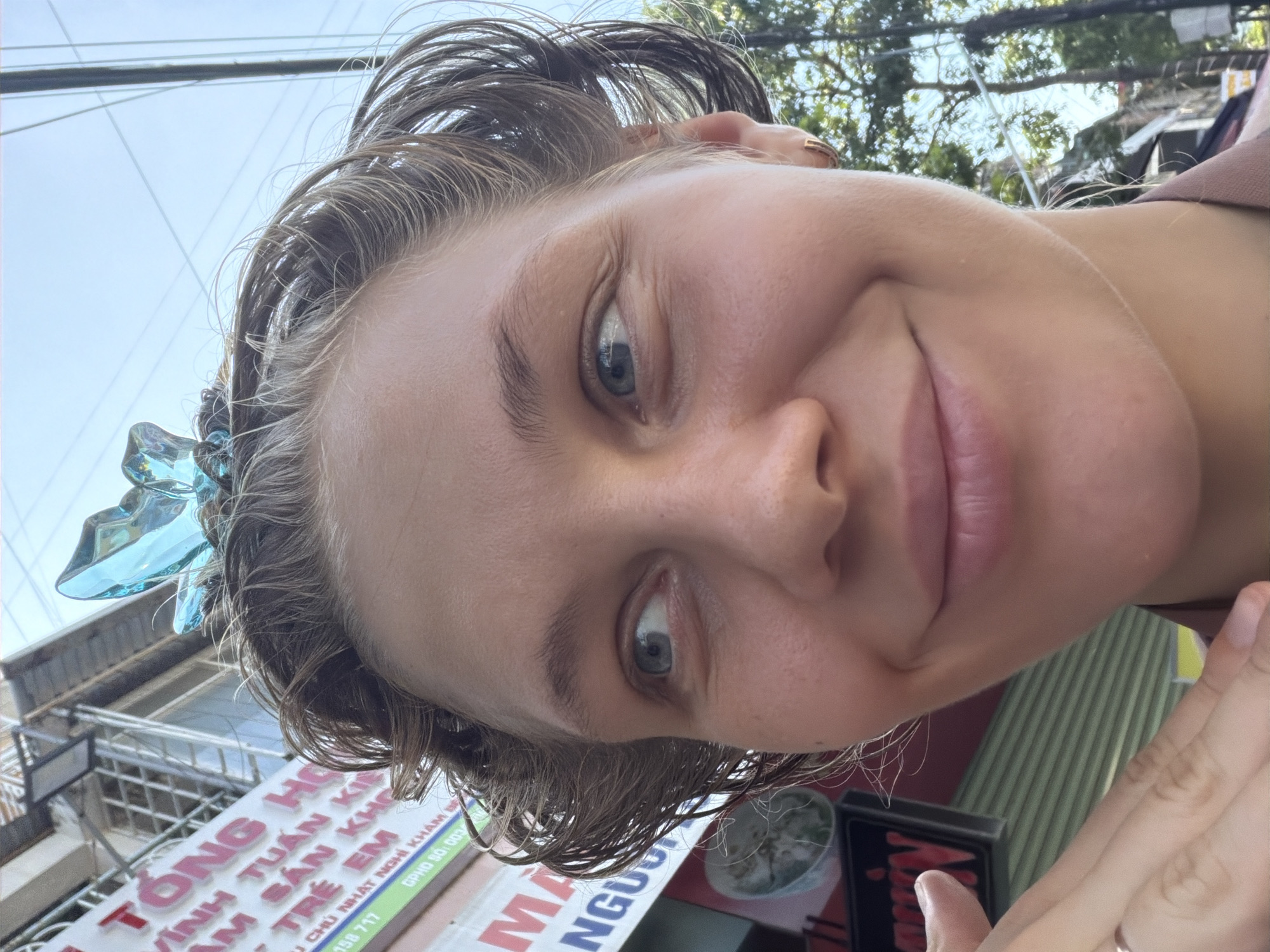 This is a close-up, tilted photograph of a young woman outdoors, with a shallow depth of field. Her face, partially obscured by her hair, dominates the frame. She has light skin, light-colored eyes, and her mouth is slightly open in a gentle smile. Her hair is wet and appears to be pulled back, with a decorative blue hair accessory visible on the left side of her head. She is wearing a light brown top with a thin strap.
In the background, the setting appears to be urban, possibly in Vietnam, given the presence of signage with Vietnamese text. The signs are for a medical facility or pharmacy, with text including "TONG HO," "TRẺ EM," "VĨNH TUẤN KIN," and "AM SẢN KHO," along with phone numbers. The time of day appears to be daytime, with bright, natural light illuminating the scene, suggesting fair weather. The overall emotion conveyed is cheerful and relaxed.