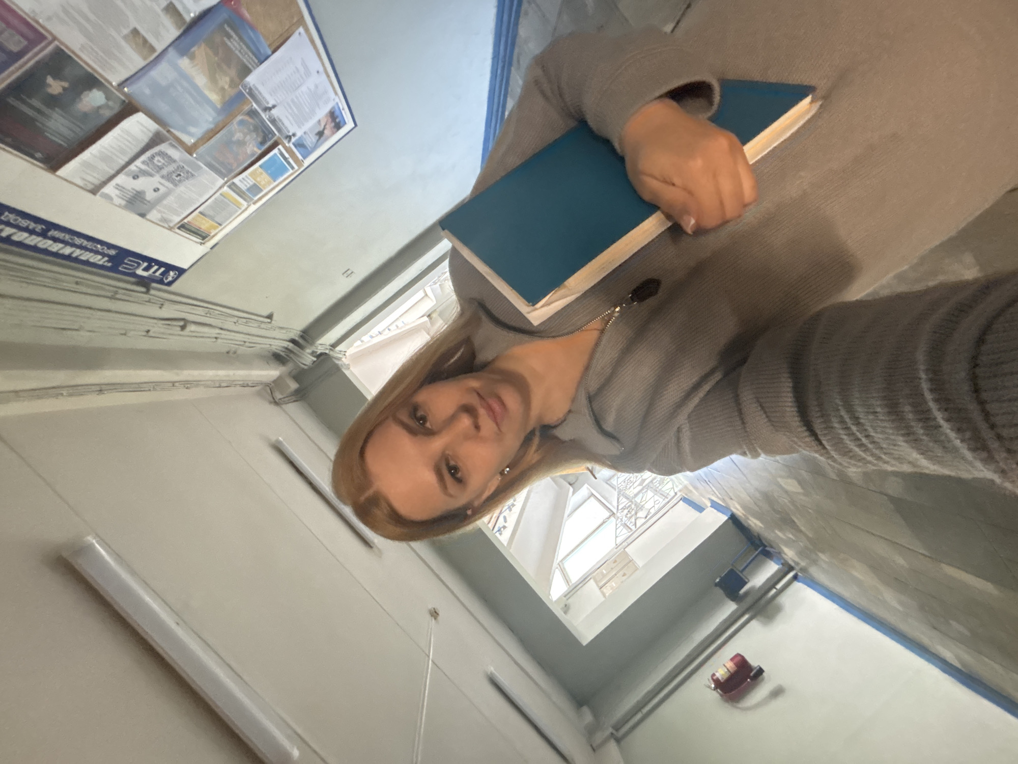 A selfie taken indoors. A blonde woman holds a blue book. The location is identifiable as being in Russia, specifically Rusanovo based on the bulletin board displaying a logo in Russian. Fluorescent lighting fixtures are visible on the walls. A fire extinguisher is located on the floor. The woman is wearing a gray top.