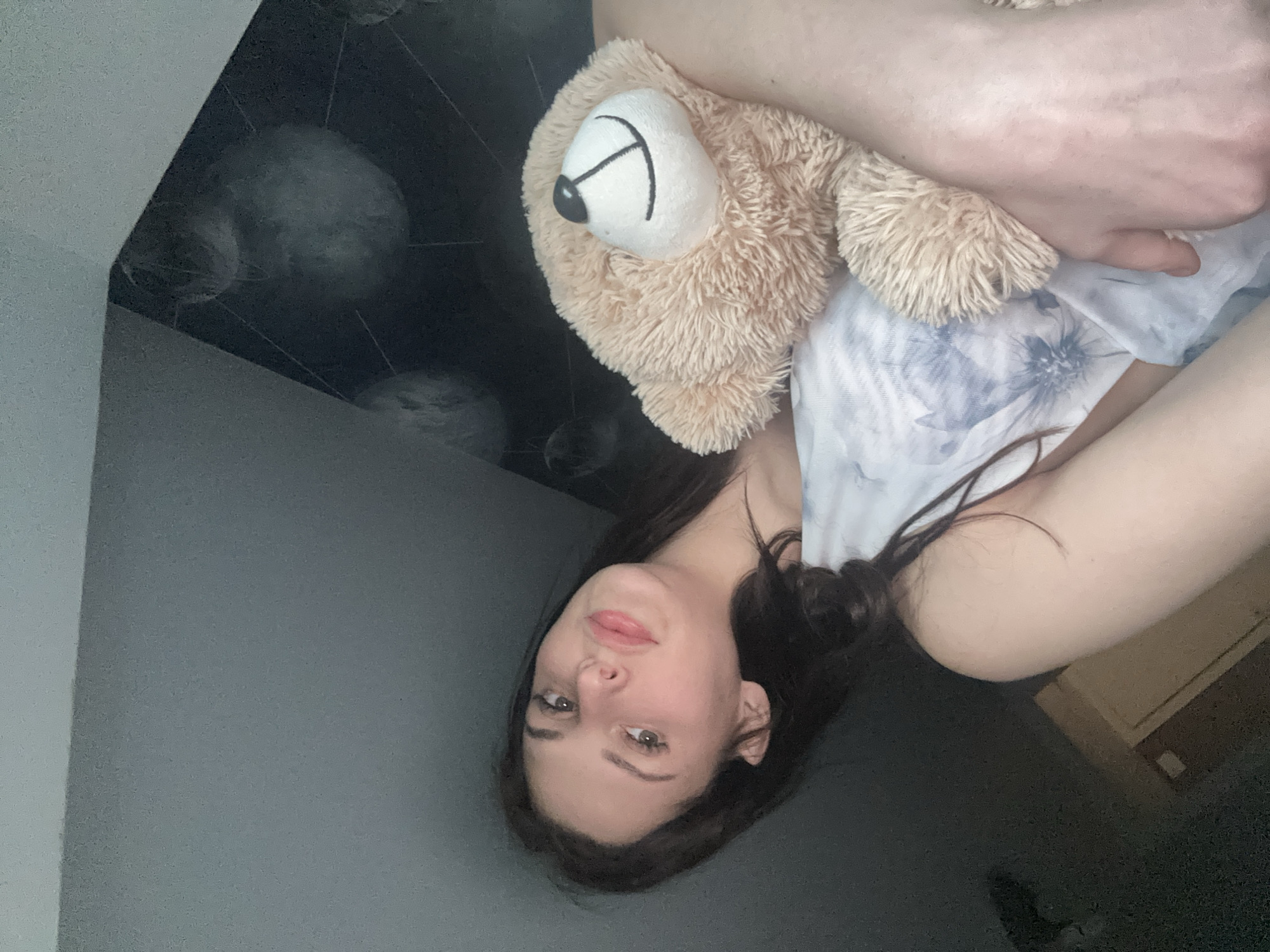 A young woman with dark hair and light skin is pictured indoors, holding a light brown teddy bear. She is wearing a light-colored garment with a blue floral pattern. Her left arm is around the bear, and her face is turned towards the camera with her head tilted. The background is dark and appears to be a wall with a circular pattern. The lighting suggests an indoor setting with limited light sources. The image is a close-up shot, focusing on the woman and the teddy bear.