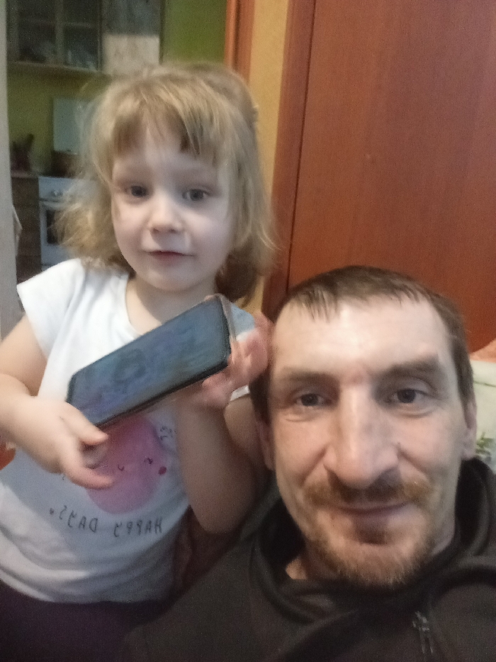 The image depicts an indoor scene in Yarun, Ukraine, featuring two individuals. On the right, a male adult with short dark hair, a mustache, and a short beard is visible from the chest up, looking directly at the camera with a slight smile. He is wearing a dark-colored hooded sweatshirt. To his left and slightly behind him is a young female child with blonde hair and bangs, also looking at the camera with a slight smile. She is wearing a white short-sleeved t-shirt with a faint pink graphic and inverted text that appears to read "HAPPY DAYS". The child holds a dark-colored smartphone horizontally in both hands, positioned between herself and the man. The background to the far left shows a blurred glimpse of what appears to be a kitchen area with shelves and light-colored walls, while the background to the right consists of a reddish-brown wall or door panel.