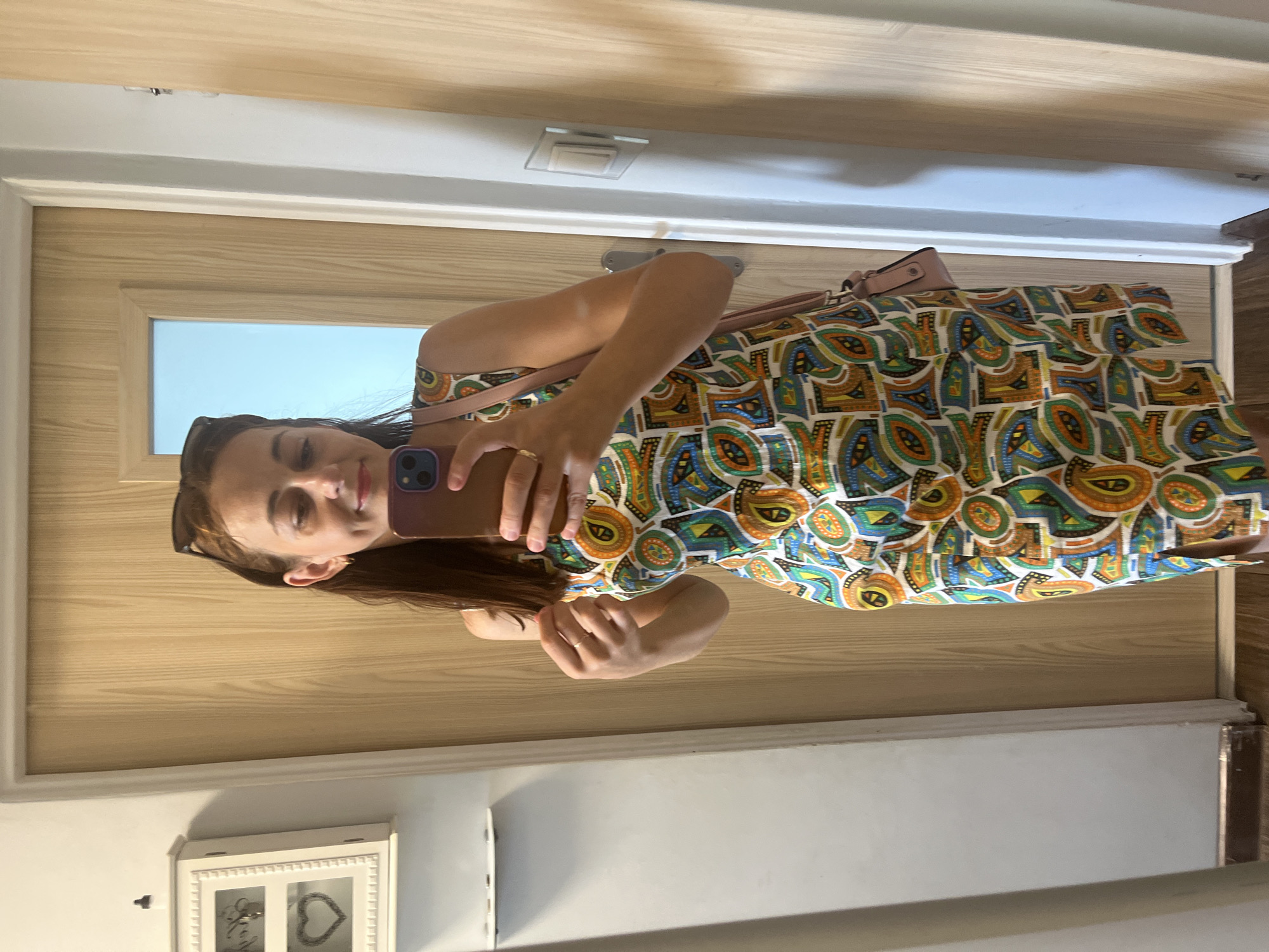 A woman is standing in front of a mirror, taking a selfie. She is wearing a colorful, patterned sleeveless dress and a light pink crossbody bag. Her hair is dark brown and she has sunglasses perched on top of her head. The background appears to be a hallway or a room with wooden doors and a white wall. There is a white cabinet with a glass door and decorative heart on the left side of the image. The floor is wooden.