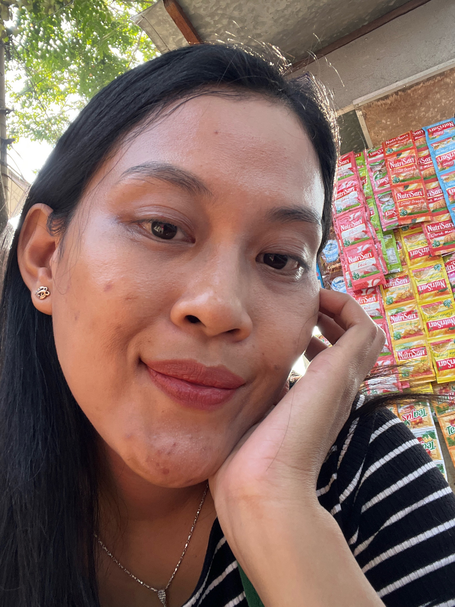 A woman with dark hair and a striped shirt is pictured in an outdoor setting. She is leaning her head on her hand, and her face is the primary focus. Behind her, a display of small packets of assorted flavors of NutriSari, a beverage mix, is visible, suggesting a market or store environment. The background also shows some greenery, indicating an outdoor location. The lighting suggests it is daytime.