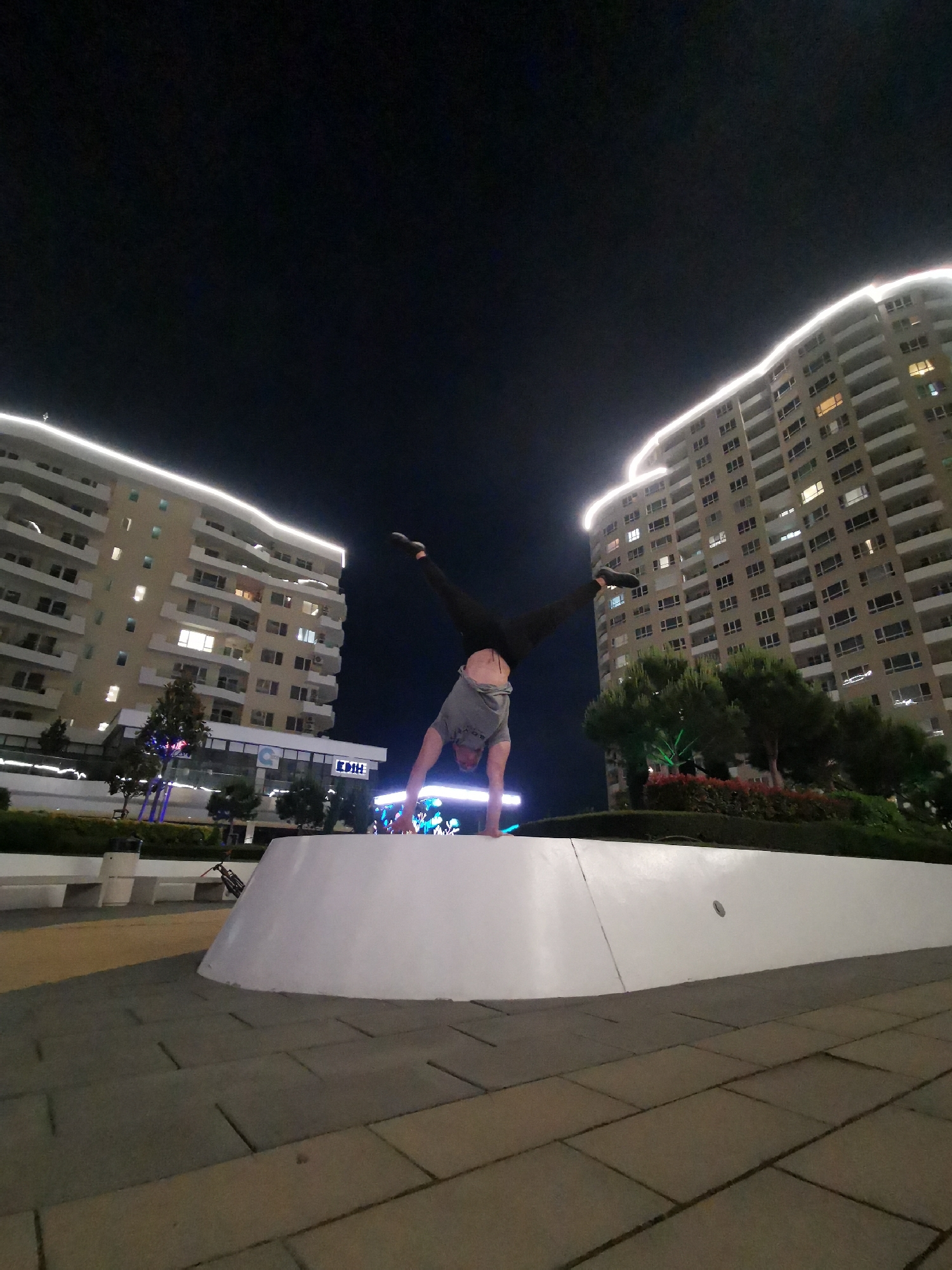 The image depicts a nighttime urban scene in Burgas, Bulgaria, centered on a male individual performing a handstand. The handstander is positioned on a low, white, smoothly curved concrete or masonry structure located in the foreground. He is wearing a grey t-shirt, dark trousers, and dark footwear, with his midriff and lower back exposed due to the t-shirt riding up. His legs are spread in an inverted "V" shape, and his hands are placed flat on the white surface, supporting his body weight.
The ground in the foreground and midground is paved with interlocking tiles of varying grey and beige tones. To the left of the central figure, a white, modern-style bench is visible, with a bicycle leaning against or near it. Further back on the left, a multi-story residential building with a light-colored facade (possibly beige or light yellow) rises prominently. This building features numerous balconies and windows, some of which are illuminated. Its perimeter, particularly the top edge and curves, is outlined with a continuous strip of bright white LED lighting. Below the residential section, a commercial ground floor is visible with signage, including a blue circular logo and text that appears to read "EDIH."
To the right of the central figure, another similar multi-story residential building stands, also featuring a light-colored facade, balconies, and illuminated windows. This building, too, is distinctively outlined with white LED lighting along its upper edges. Between the central figure and the right building, a landscaped area with bushes and small trees is visible, some of which are illuminated with colored lights, including green.
The sky above is dark and cloudless, indicating night. The overall scene is well-lit by artificial sources from the buildings and street-level installations, highlighting the modern architectural style characteristic of newer developments in cities like Burgas, particularly in residential and commercial districts.