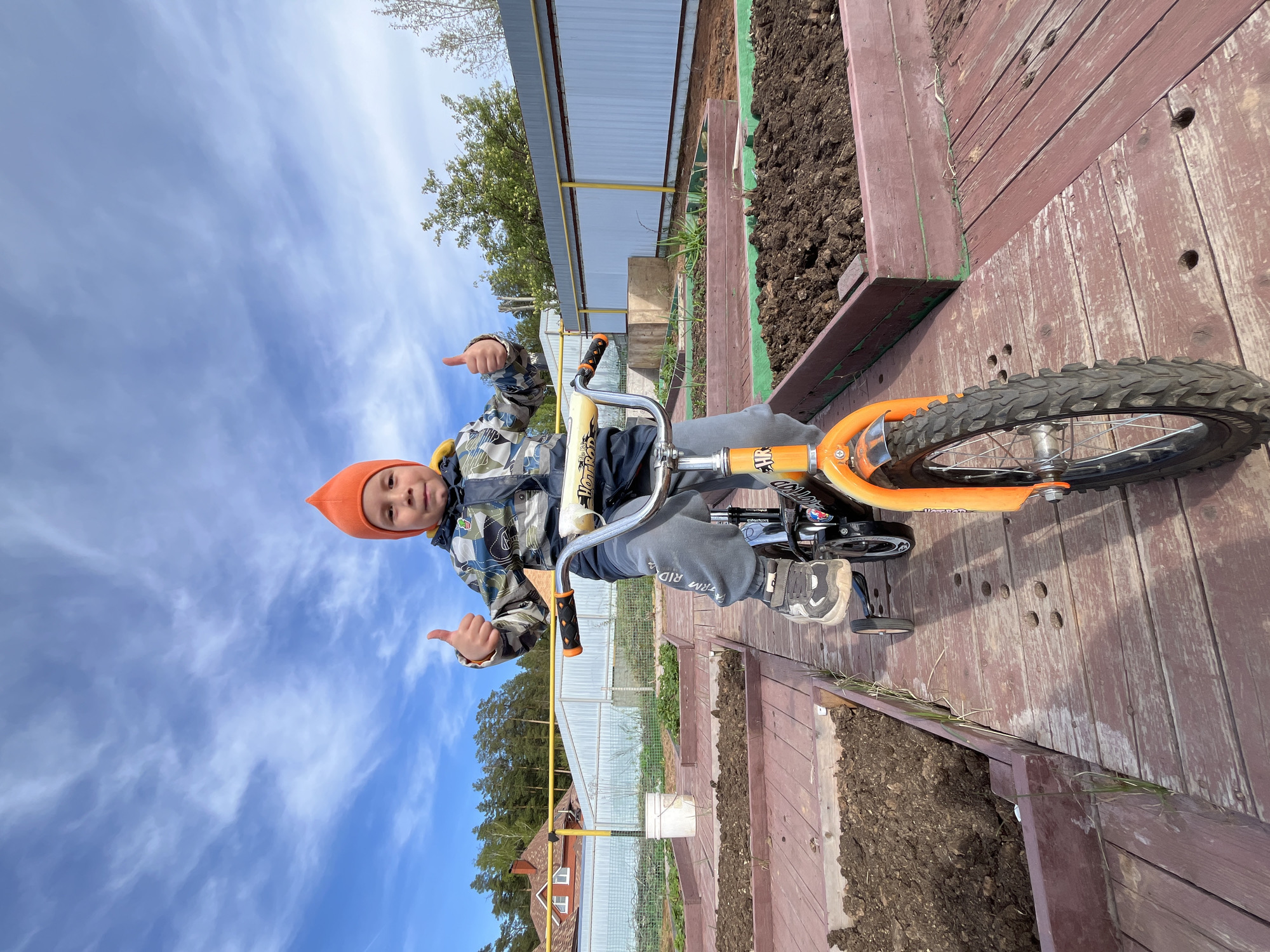 A young boy wearing an orange knit cap and a camouflage jacket sits on an orange bicycle, giving two thumbs up to the camera. The background consists of a blue sky with streaks of clouds, a metal fence, and a greenhouse. He is positioned on a wooden platform bordered by garden beds filled with soil. The word "RIDE" is visible on the boy's sweatpants. The location may be Chaikovsky, Russia.