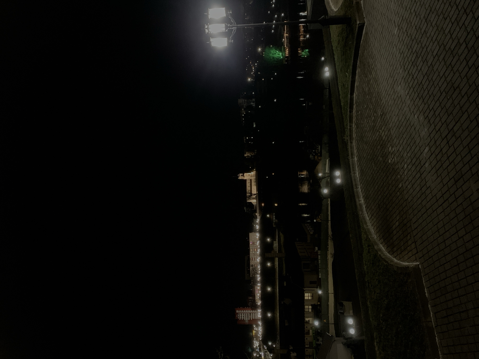 The image depicts a night scene in Hrodna, Belarus, showing a waterfront promenade along the Neman River. A brick-paved walkway is on the right side of the image, illuminated by spaced streetlights. To the left, across a dark expanse, is the city skyline, marked by building lights. A distinctive cluster of green lights is present, potentially indicating a park or recreational area. The scene is dark with artificial lighting playing a key role in illuminating the environment.