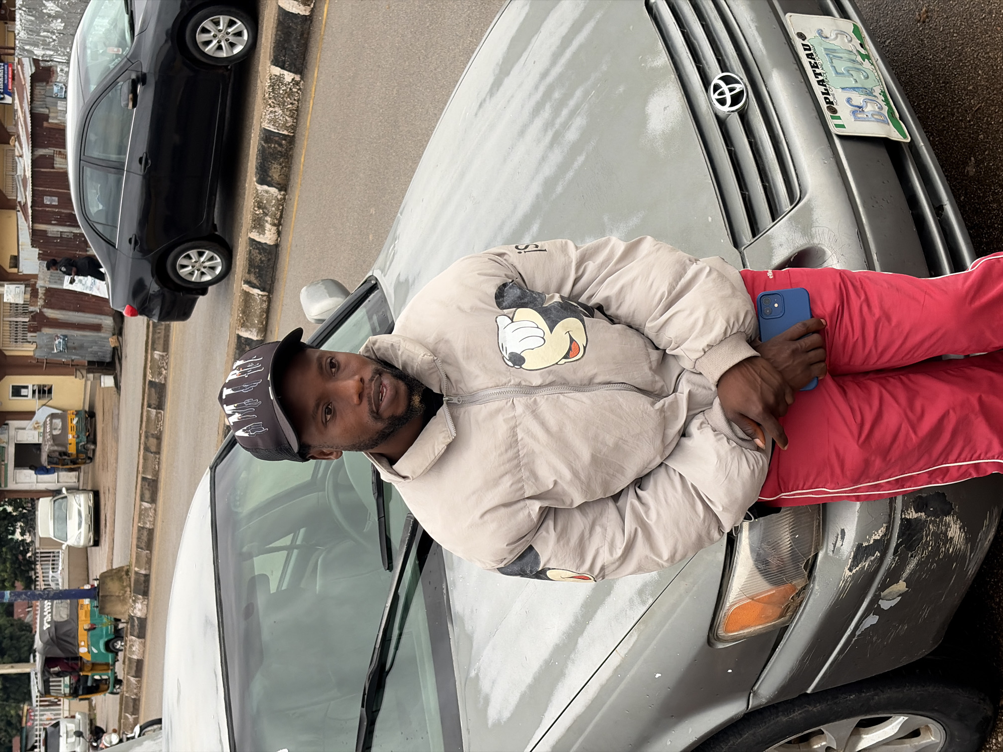 A man with a goatee and a black cap with a white design stands in front of a silver Toyota Camry in Jos, Nigeria. He is wearing a light-colored puffer jacket with a Mickey Mouse graphic on the left sleeve and red track pants with white stripes. He holds a blue smartphone in his right hand. The car is parked on an asphalt road with a curb and sidewalk visible. In the background, other vehicles, including a yellow and green auto-rickshaw, a white sedan, and a motorcycle, are present along with buildings and streetlights. The car's front bumper and headlight are damaged. A Nigerian license plate, "IIOPLATEAU BSA-571-LS," is attached to the front of the car.