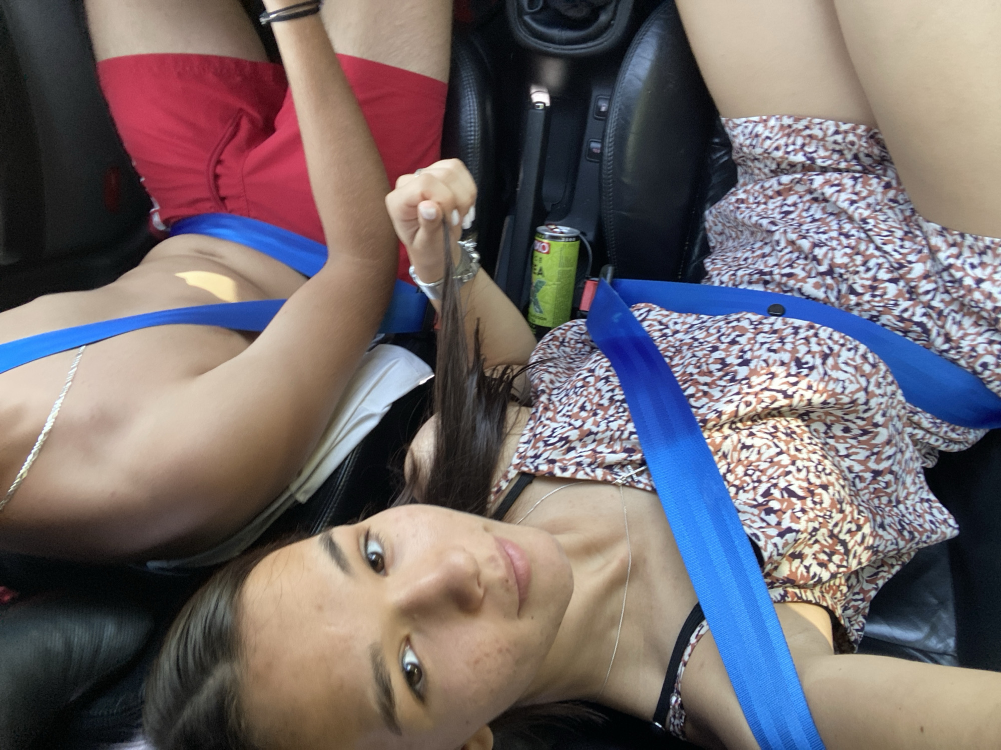 In this close-up, high-angle shot taken in Berovo, Bulgaria, two individuals, a woman and a man, are seated in a vehicle, likely on a road trip. The woman is in the foreground, her face partially visible as she looks towards the camera. She has long, dark hair and is wearing a patterned dress and a silver necklace. A blue seatbelt is prominently displayed across her chest.
Beside her, the man is seen from the upper body, wearing red shorts. He is also wearing a blue seatbelt, and his left arm rests on the woman's arm. A can of what appears to be a beverage is placed in the car's console. The interior of the car, with its black leather seats and central console, is visible. The lighting suggests it is daytime, possibly sunny, with shadows cast within the vehicle. The background is blurred, indicating a focus on the individuals and the immediate car interior.