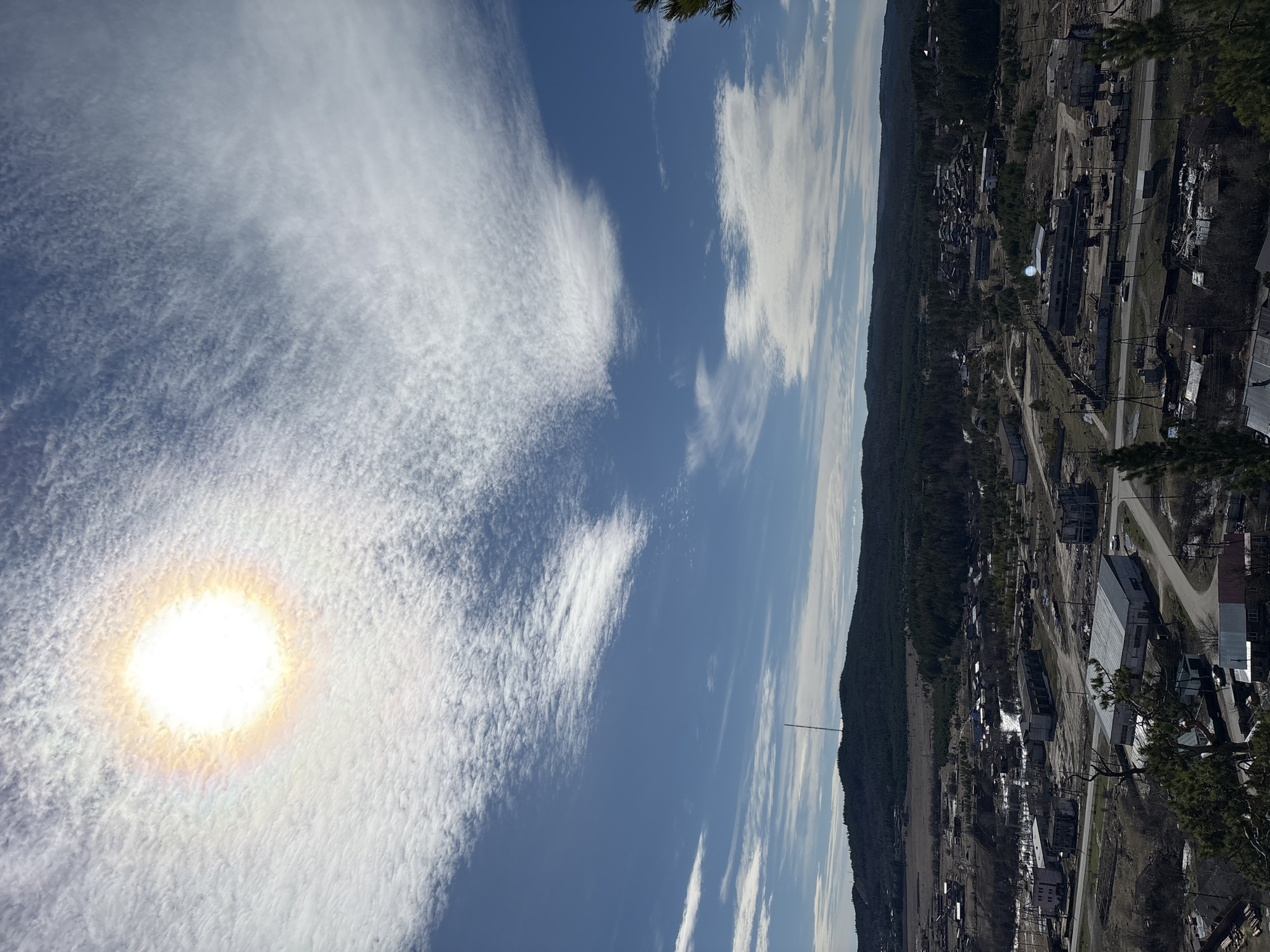 The image shows a view of the city of Невон, Russia, under a bright, sunlit sky with cirrus clouds. The sun is visible through a layer of thin clouds, creating a halo effect. The landscape includes a mix of trees and low-lying hills along the skyline, with a transmission tower visible. The urban area appears to be a small town or industrial settlement with various buildings and roads. The photo is taken from an elevated perspective, possibly a hillside overlooking the town. There are no people immediately visible in the image.