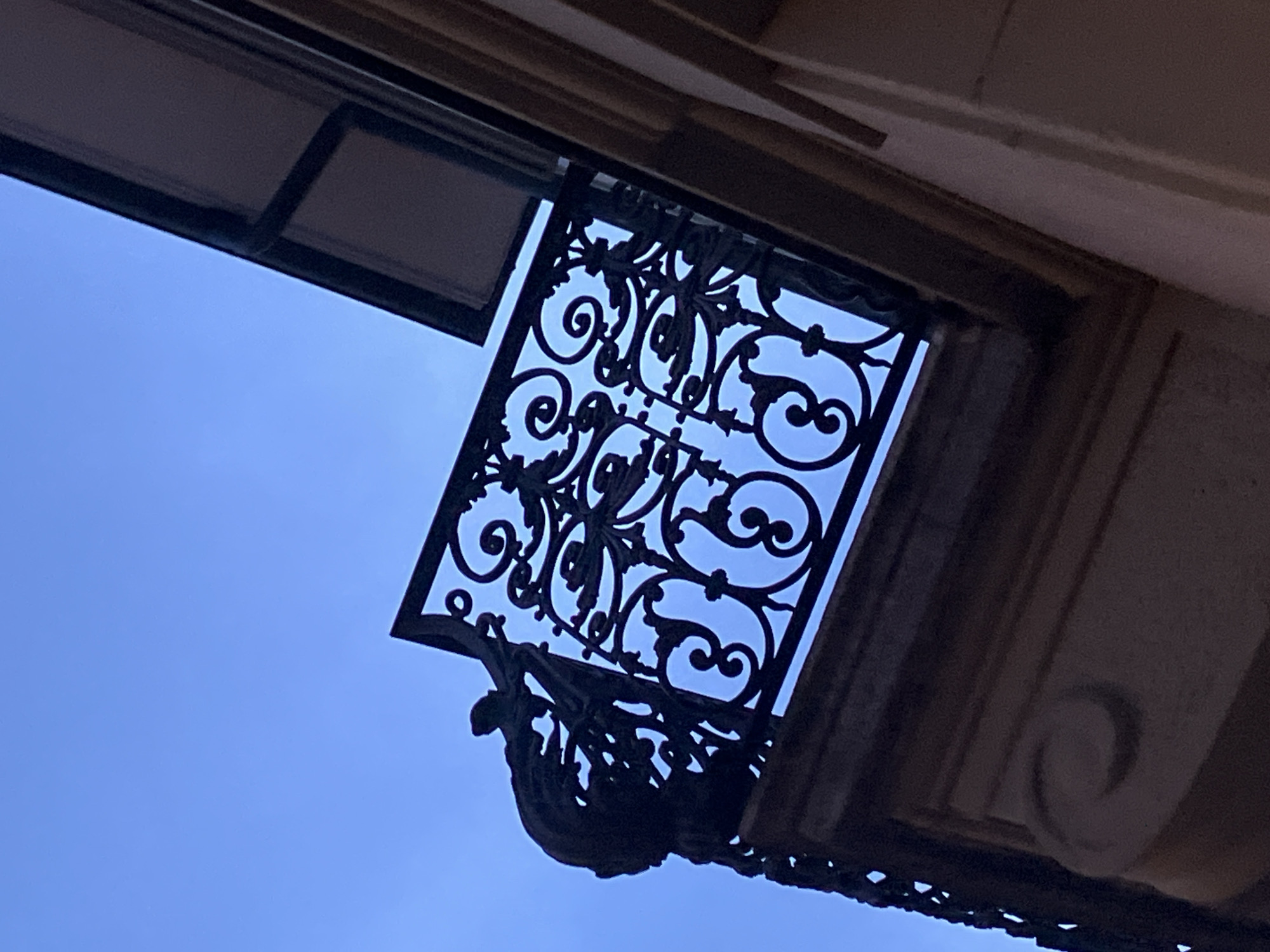 A low-angle shot captures an ornate, dark-colored iron balcony against a blue sky. The balcony is adorned with intricate patterns of swirling designs and floral motifs. The balcony is attached to a building with a stone facade. The sky is a uniform pale blue, providing a stark backdrop to the dark ironwork. Location: Unknown, Unknown.