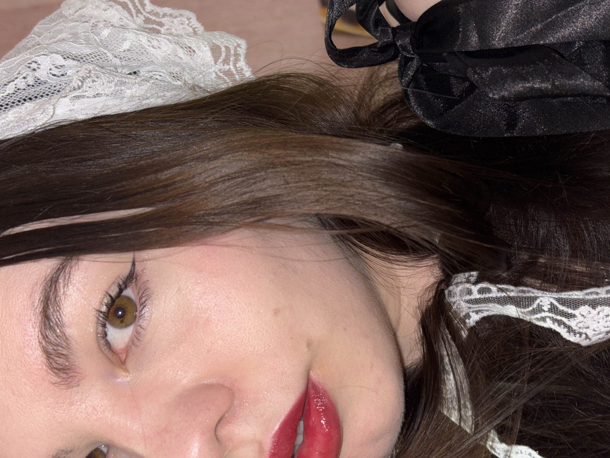 A close-up shot focuses on a person's face, positioned diagonally in the frame. The individual has fair skin, dark brown hair, and light brown eyes with black eyeliner. The lips are coated in glossy red lipstick. A white lace headdress adorns the head, partially covering the hair. A black bow is positioned near the top right corner of the frame. The background is blurred, providing minimal contextual information. The image is not related to the city of Perm, Russia.