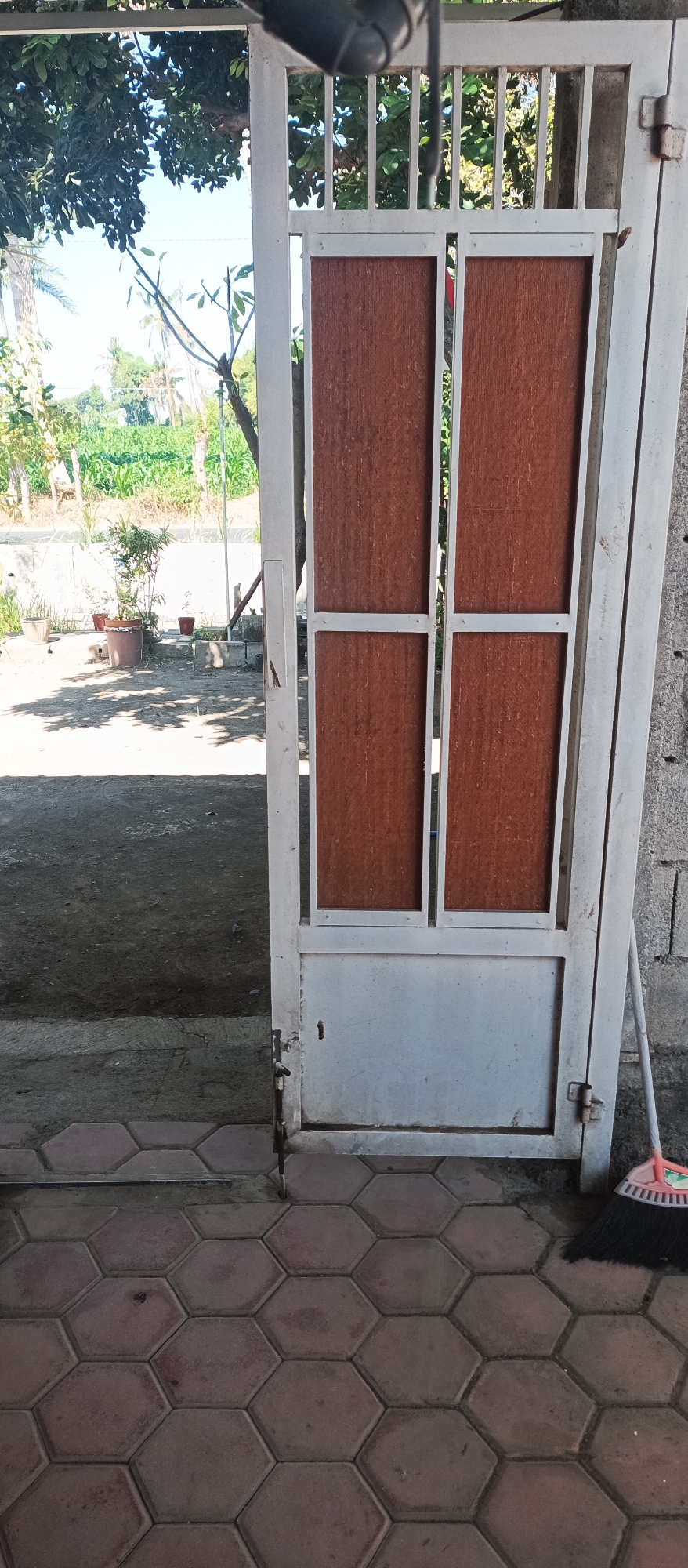 The image shows an open metal gate with brown panels, likely in Arjasa, Indonesia. The gate is painted white and has a handle and hinges visible. Beyond the gate, there is a dirt path and some potted plants. In the background, there is greenery, suggesting an outdoor setting with vegetation. A broom with pink bristles rests against the wall to the right of the gate. The ground in front of the gate is paved with hexagonal tiles. The overall scene depicts a simple entrance to a property in a semi-rural or residential area.