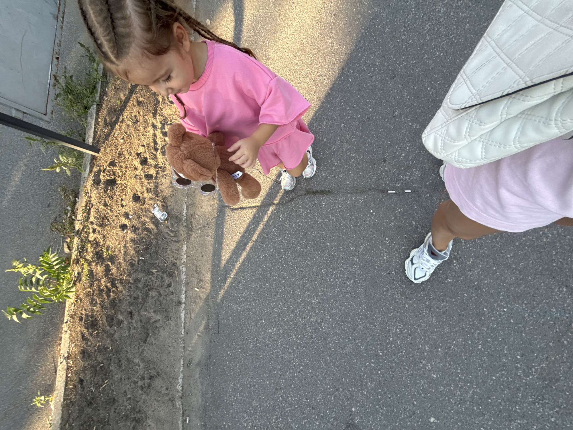 A young girl with braided hair is standing on an asphalt surface, holding a brown teddy bear. She is wearing a pink dress and white sneakers. In the foreground, a person's legs are visible, also wearing white sneakers and a pink skirt, holding a white quilted bag. The background shows a dirt patch with some plants and a discarded plastic bottle. The scene appears to be outdoors, possibly in a park or on a sidewalk in the city of Chișinău, Moldova, judging by the architecture and vegetation. The lighting suggests it is daytime, with shadows cast by the sun.