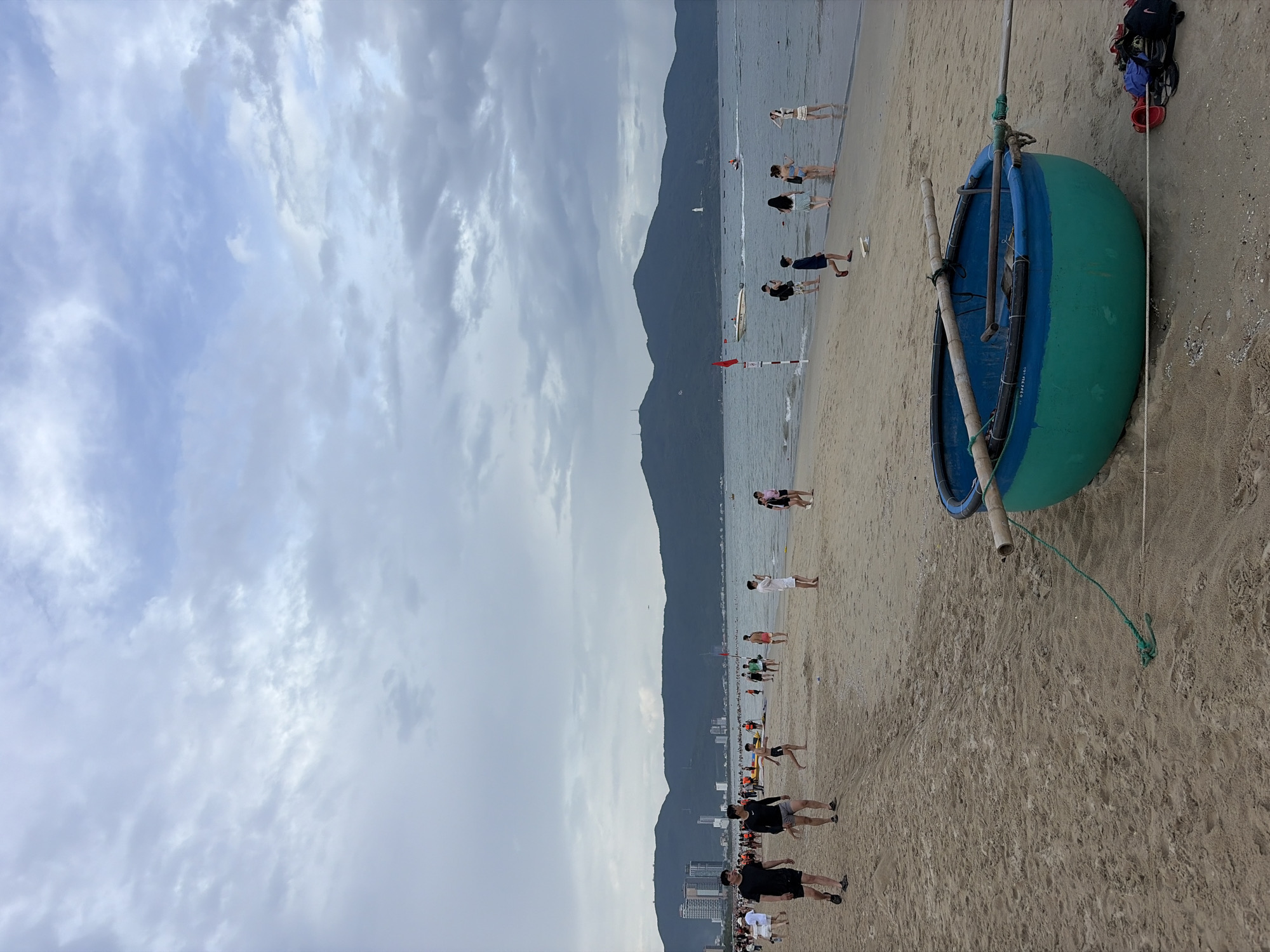 The image depicts a beach scene in Đà Nẵng, Vietnam, likely My Khe Beach, with the Marble Mountains visible in the background. The sky is overcast with scattered clouds, suggesting either early morning or late afternoon. In the foreground, a traditional Vietnamese basket boat, painted in vibrant green and blue, rests on the sand. A wooden oar extends from the boat, tied with a green rope. Several people are scattered across the sandy beach, some walking, some standing near the water's edge, and some appearing to be engaged in various activities. The sea is calm with a few boats visible in the distance, including a white motorboat and a jet ski. Along the shoreline, there are buildings and the distinctive silhouette of the Marble Mountains, a prominent landmark of Đà Nẵng. The overall atmosphere is one of a relaxed beach day with both locals and tourists enjoying the coastal environment.