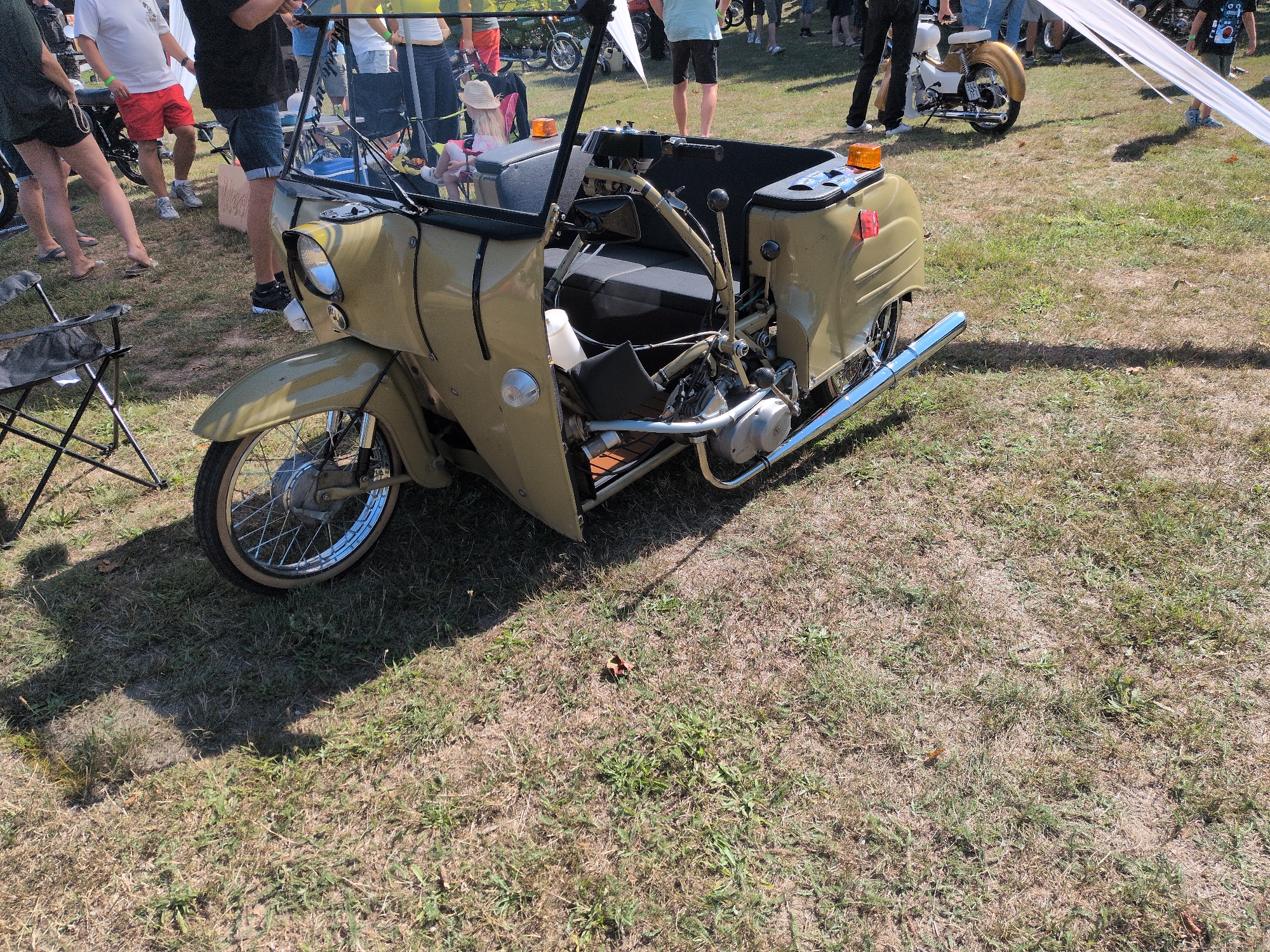 The image depicts an outdoor gathering, possibly a market or event, on a grassy area. A vintage tan three-wheeled scooter with a sidecar is prominently featured in the foreground. The scooter has a single front wheel and two rear wheels, with a cargo area behind the driver's seat. Several people are visible in the background, some standing and some sitting. The environment is characterized by dry grass and scattered patches of green. The lighting suggests it is a sunny day. The scene appears to be set in a rural or park-like environment, consistent with the provided location of Hohenferchesar, Germany, which is a municipality in the Havelland district of Brandenburg.