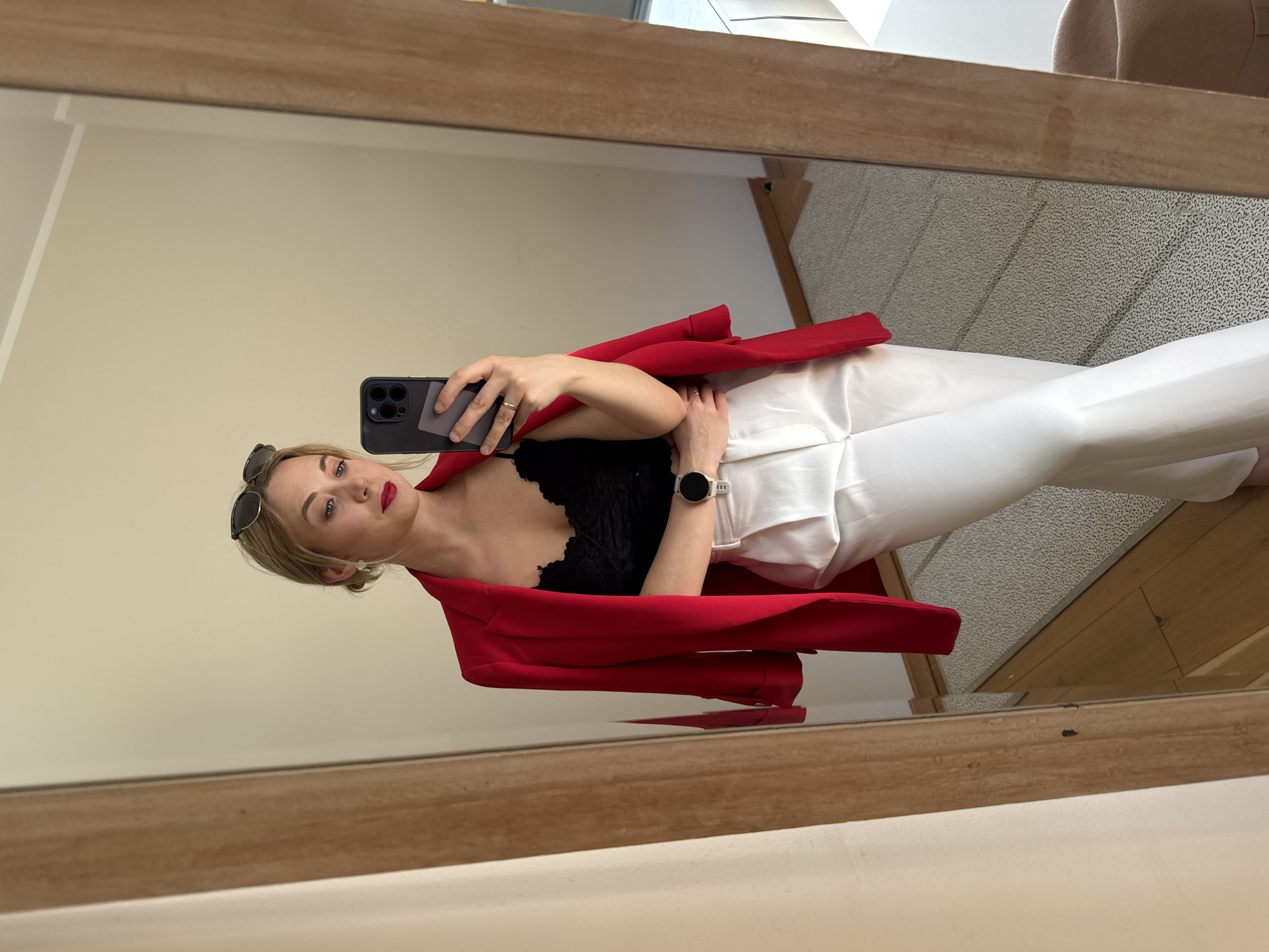 A woman takes a mirror selfie in an indoor setting, likely in Riga, Latvia. She is reflected in a large mirror with a prominent wooden frame. The woman has blonde hair, blue eyes, and is wearing red lipstick. Sunglasses are perched on her head.
She is dressed in a vibrant red blazer draped over her shoulders, a black lace top or bralette, and high-waisted, wide-leg white trousers. On her left wrist, she wears a white smartwatch, and rings are visible on her left hand. She holds a dark-cased smartphone with a gray attachment in her right hand, which partially covers her face as she angles for the photo.
The background reflected in the mirror shows a plain, light-colored wall and a light-toned carpet with a subtle pattern. Wooden elements, possibly part of the floor or furniture, are also visible. A beige upholstered chair is partially seen in the upper right corner of the reflection. The lighting is bright and even, indicating daytime. A small white tag or label is visible on the waistband of her white trousers, but any text on it is not discernible. No other visible text is clearly legible in the image.