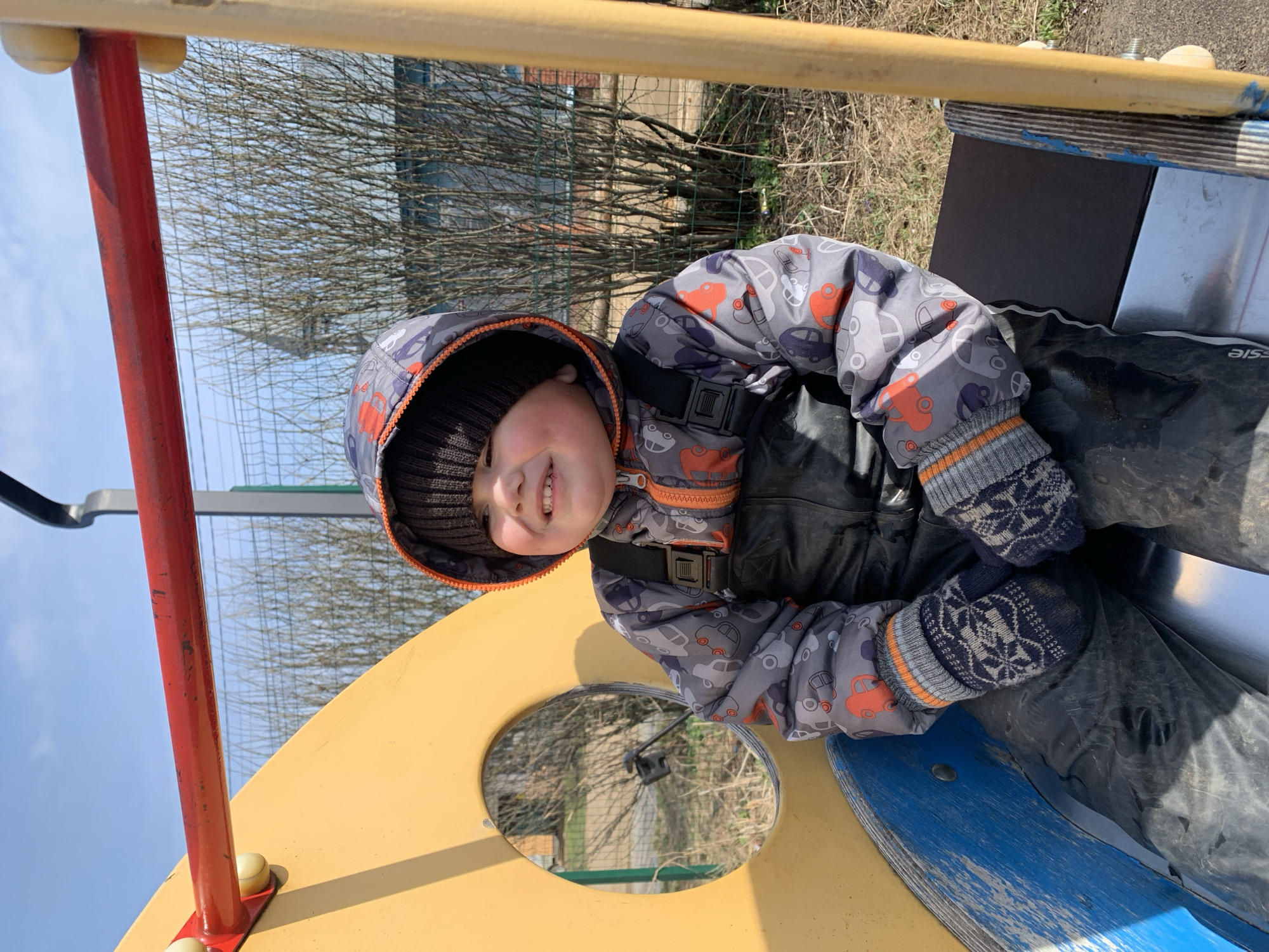 A child wearing a patterned grey jacket and knitted hat is sitting on a yellow and blue playground structure. The structure also has red and yellow poles. The child is smiling, and their face is slightly turned to the left. They are wearing mittens with a snowflake pattern. The background includes bare trees, a fence, and a building. The location is Фоки, Russia.