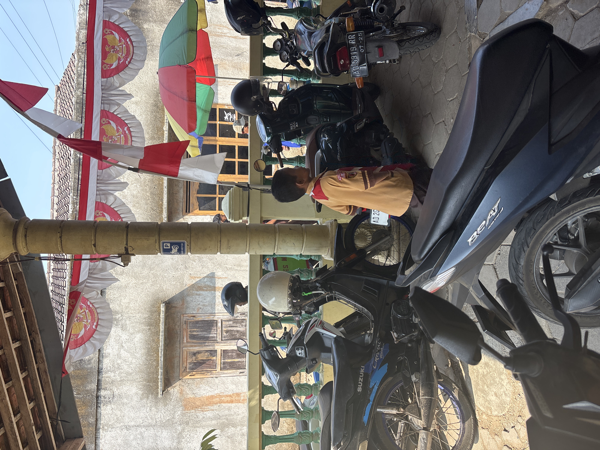 The image depicts an outdoor scene with multiple motorcycles parked under a shaded structure, likely a carport or veranda. A young boy, wearing a light brown and red school uniform, stands facing to the left, looking away from the camera. He is positioned near a dark green scooter. Behind the boy, several motorcycles are visible, including a black Suzuki and a Honda with a license plate reading "AD 5319 RR". Red and green umbrellas are open and positioned behind the motorcycles. The background shows a building with a textured, light-colored wall and wooden-framed windows. Decorative red and white banners with a central emblem are hung along the roofline of the building and the veranda. The ground is paved with interlocking hexagonal stones. The overall impression is a mundane, everyday scene in what appears to be a residential or community area in Tirtomoyo, Indonesia, possibly decorated for a local event or holiday given the prominent Indonesian flags and banners.
