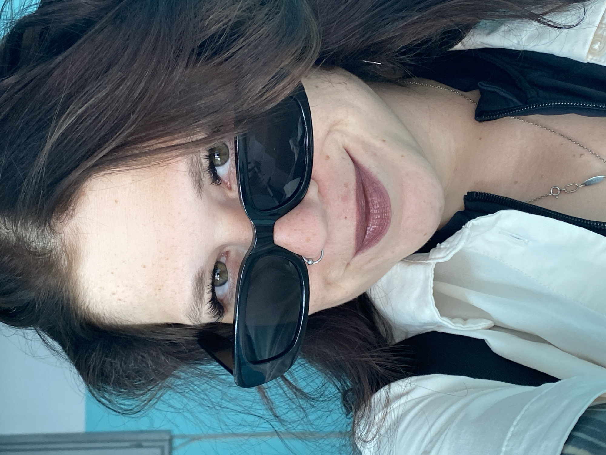 A close-up, low-angle, selfie-style photograph shows a person with dark brown hair, light skin with freckles, and green eyes. The person is wearing large, black, rectangular sunglasses, a white collared shirt, and a black jacket. They have a nose ring and are smiling. The background is a plain turquoise wall. The image appears to have been taken indoors. The person is likely a resident of the city of Chaikovsky, Russia.