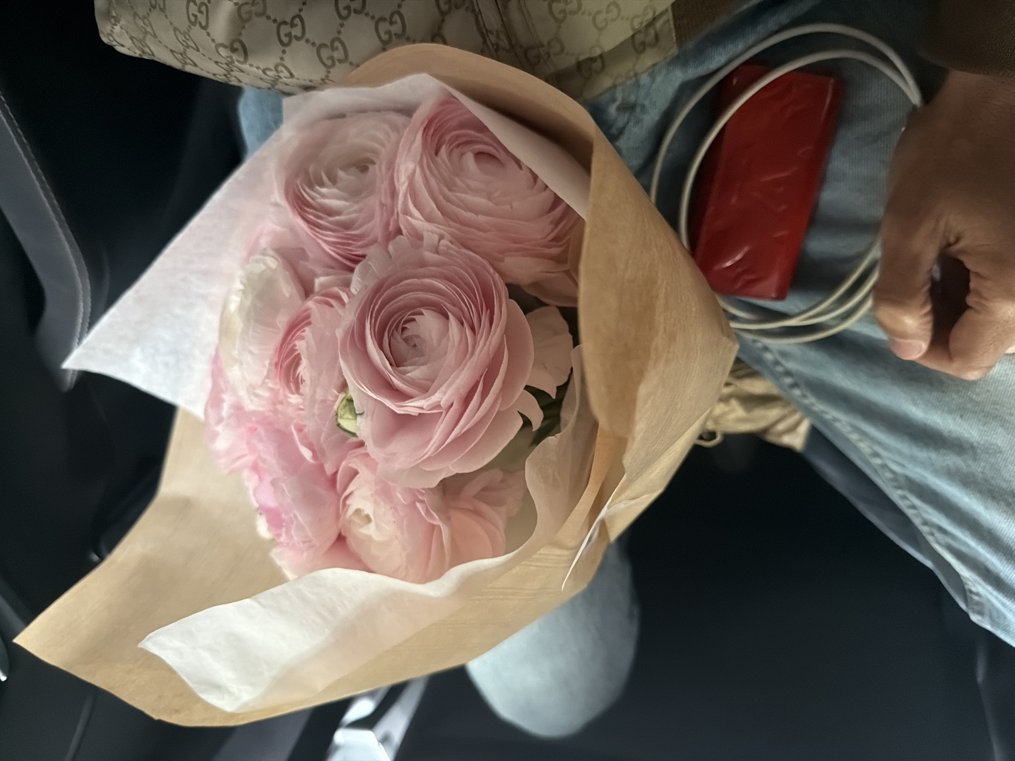 The image shows a bouquet of pink ranunculus flowers wrapped in brown paper. The bouquet is situated next to a person wearing blue jeans and holding a red rectangular object with white cables. A Gucci purse is visible in the background. The location is unknown, there is no information in the image to place the scene in Paris, France.