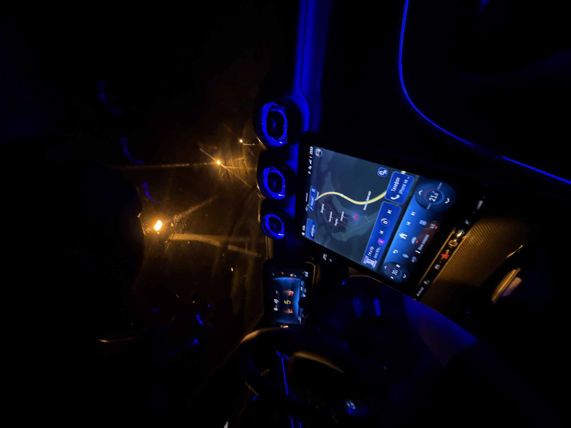 The media file captures the interior of a modern car at night, illuminated by blue ambient lighting and digital displays. The perspective is from within the vehicle, focusing on the dashboard and partially revealing the exterior through the windshield.
A large, central infotainment screen dominates the right side of the frame, displaying a navigation map with a yellow route line. It also shows media playback details ("I'm a VIP - Artist: Wh..."), a phone interface with Cyrillic text "Телефон iPhone 11s...", and climate control set to "21.6°". The top right corner of this screen indicates "2:59 PM" and "100 km/h". To the left of the main screen, three prominent circular air vents glow with blue light, mirroring the continuous blue light strip along the upper dashboard.
A smaller digital display behind the steering wheel shows "P" and the number "7". The steering wheel itself is partially visible on the bottom left, also featuring blue accents. Through the windshield, several blurred, warm-toned streetlights are visible, confirming the nighttime setting. There are reflections on the windshield, possibly from interior lights or external moisture. A person's arm and shoulder are faintly visible on the far left. The overall impression is a sleek, contemporary car interior, possibly in motion or parked in Zvezditsa, Bulgaria.