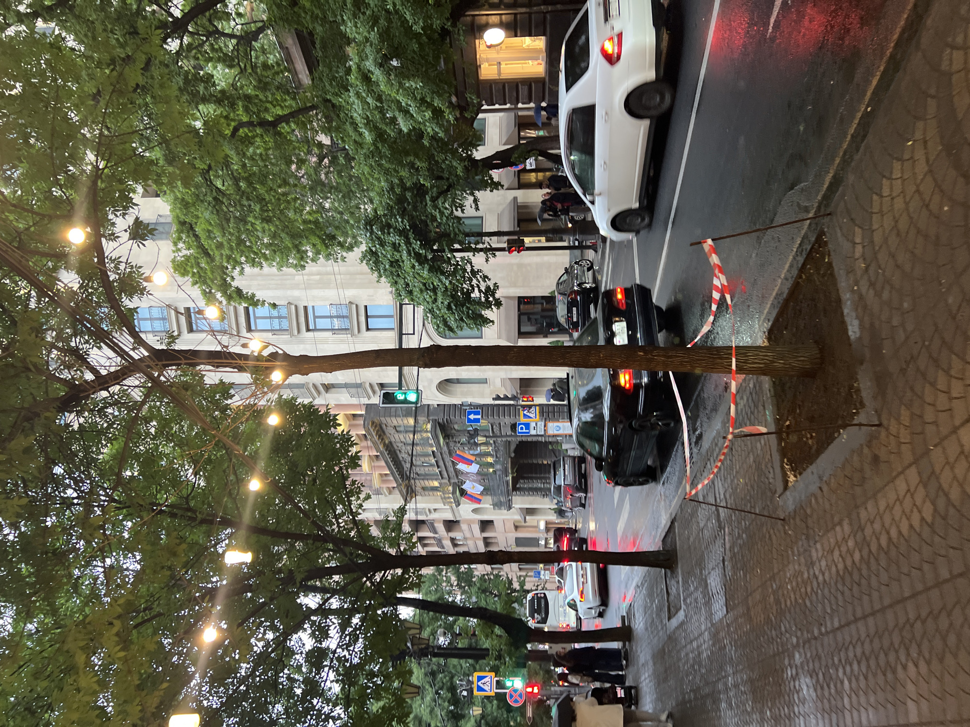 The image depicts a Yerevan, Armenia street scene at dusk or night. A tree with string lights is in the foreground along the sidewalk. The sidewalk is made of cobblestone. A damaged tree is being held up with poles, and wrapped with red and white caution tape. The street is wet from rain. Cars, including a white vehicle and a black vehicle, are on the street. Buildings line the street, with flags displayed from one. Traffic signals and street signs are visible. Pedestrians are walking along the sidewalk.