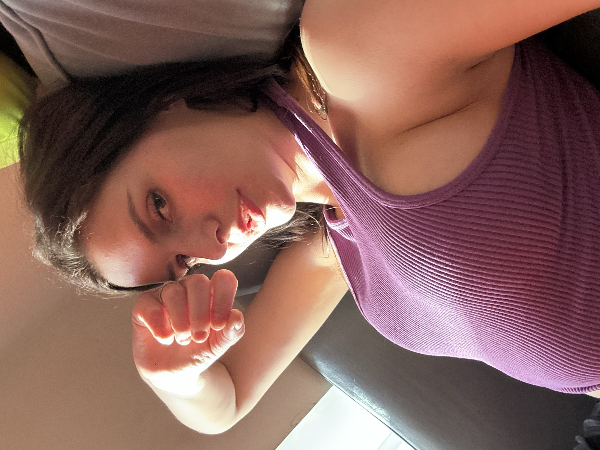 A young woman with dark hair is shown in a close-up, posed shot. She is wearing a ribbed, purple tank top. Her right arm is bent upwards, with her hand near her face, obscuring her forehead. Her left hand is resting on her chest. The background is blurred, but appears to be indoors, possibly a sofa or chair. Sunlight illuminates her face and arm, creating highlights and shadows. Her lips are slightly parted, and her gaze is directed towards the camera.