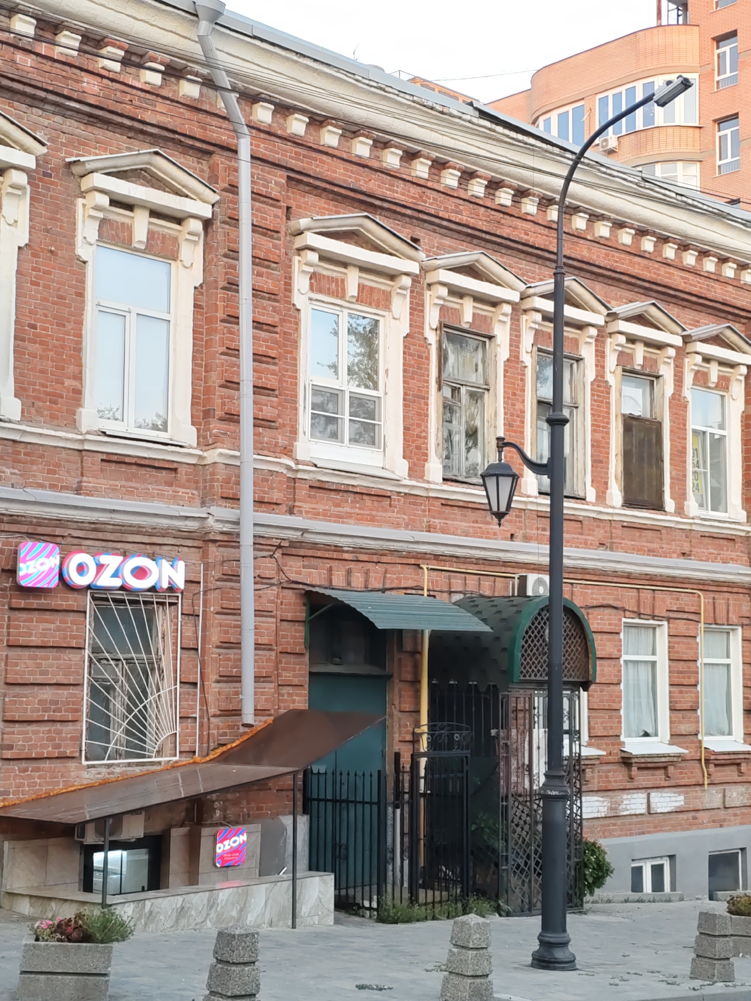 The image shows a brick building with several windows. A street lamp is in the foreground. On the left side of the building, there is a sign that says "OZON". There are also some potted plants and decorative stones on the sidewalk in front of the building. In the background, another building with a curved facade and many windows is visible. The sky is overcast. The overall scene depicts a street in a city.