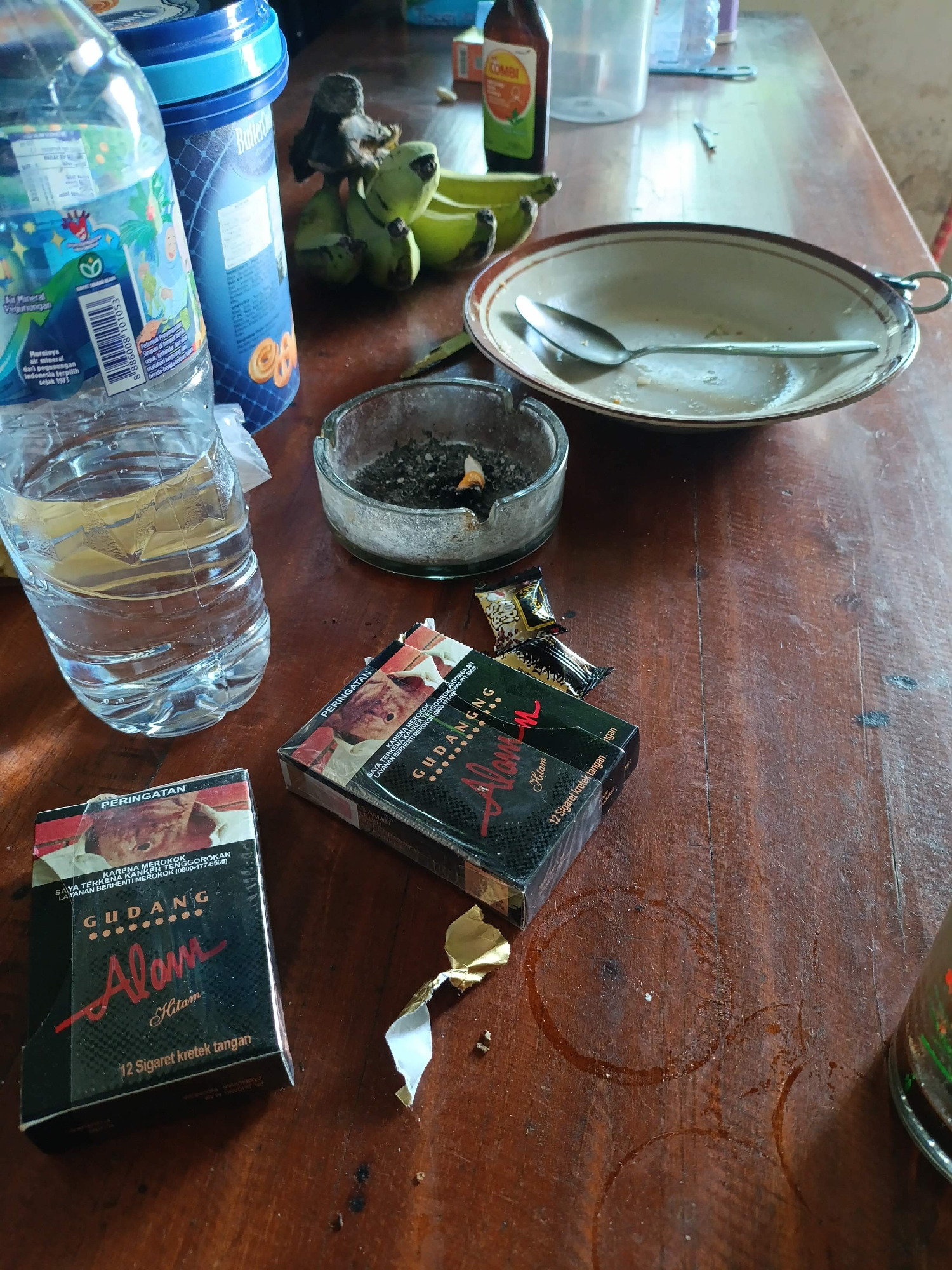 A close-up, high-angle shot captures a wooden table with various objects, suggesting a moment of relaxation or a break. In the foreground, two packs of "Gudang Alam" cigarettes, a brand commonly found in Indonesia, are placed side by side. One pack is open, revealing a glimpse of the cigarettes within. Next to the cigarettes is a partially unwrapped candy or confection, its gold foil wrapper glinting. To the left, a clear plastic bottle of water stands, its contents reflecting the ambient light. In the background, a glass ashtray is filled with cigarette butts and ash, with one cigarette still smoldering, indicating recent use. A spoon rests in a shallow ceramic bowl, with remnants of food or residue inside. A bunch of green bananas is also visible, alongside a bottle of what appears to be a medicinal tonic, possibly "Combi." The wooden table shows signs of use, with water rings and minor scuffs. The overall scene evokes a casual setting, possibly a home or a simple establishment in Adipala, Indonesia.