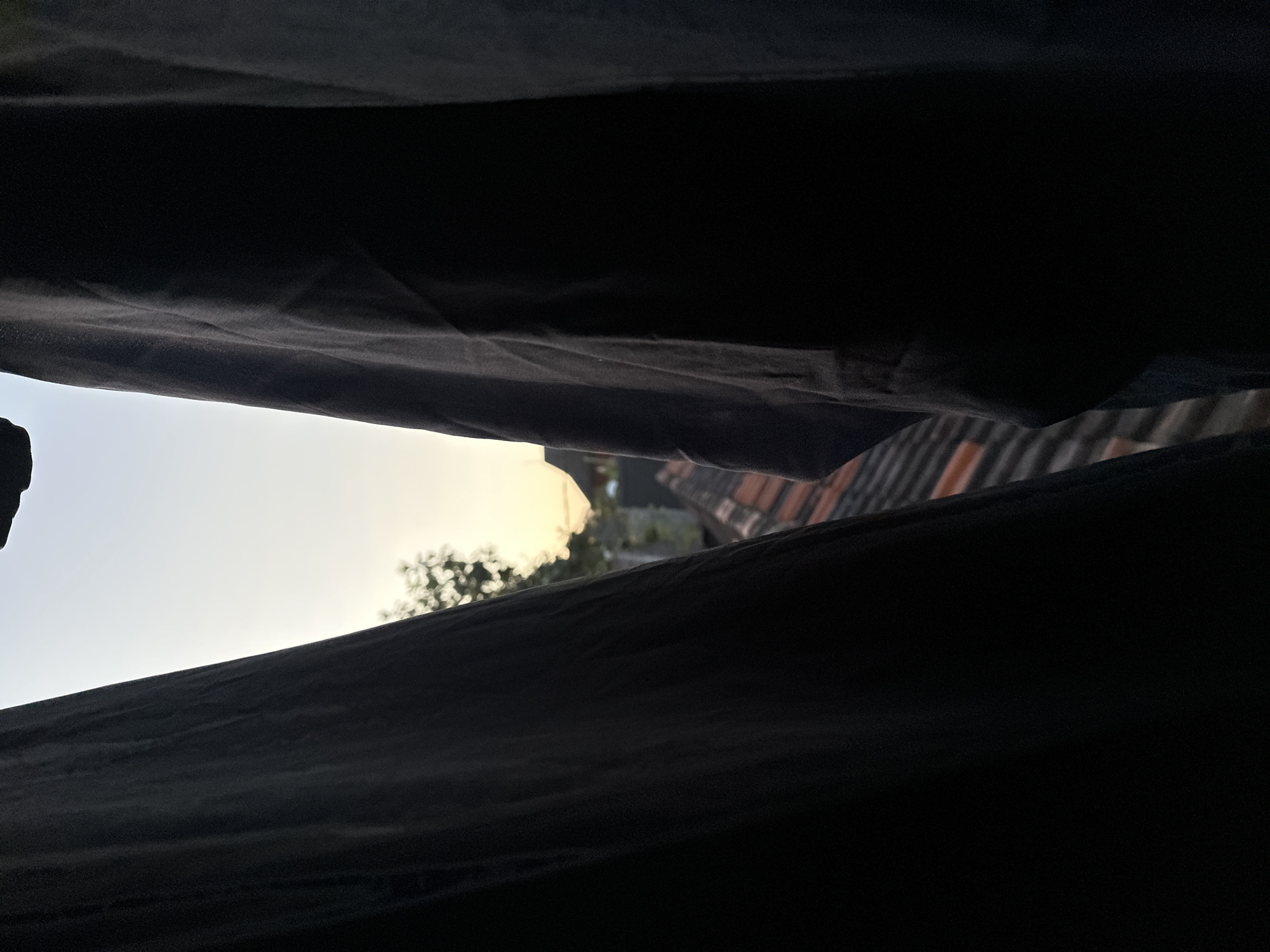 The image depicts a view from behind dark curtains, looking out at a twilight sky with a hint of sunset colors. The foreground is dominated by the dark fabric of the curtains, which obscure most of the scene. Peeking through the gap between the curtains, one can see a rooftop with orange tiles and some greenery. In the distance, faint outlines of buildings are visible against the pale sky. The overall impression is one of a quiet evening, possibly in a residential area. The specific location in Bulakrejo, Indonesia, is not explicitly evident from the visual cues alone, but the scene suggests a domestic setting with a view of the sky and surrounding rooftops.
