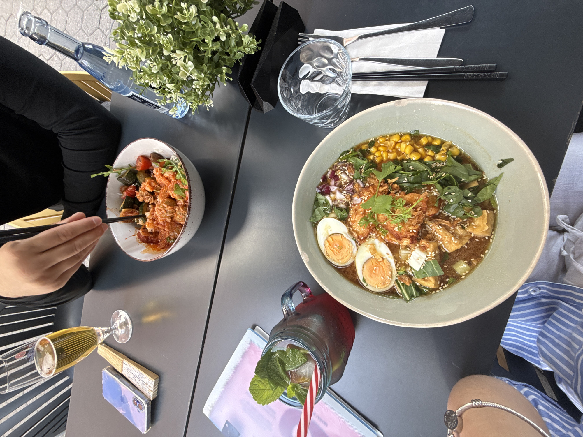 The scene is a top-down view of a table at a restaurant, likely outdoors. The location is indicated to be Aschaffenburg, Germany, though this is not visually evident in the photograph. Two people are present, one on the left wearing black clothing and using chopsticks to eat from a bowl of pasta, and another on the right wearing a blue and white striped shirt and a silver bracelet. On the table, there are two bowls of food: one with pasta and the other with ramen containing corn, eggs, and greens. Additionally, there is a bottle of water, a glass, cutlery and chopsticks, a flower pot, a glass of champagne, a smartphone with a fan on top, and a mason jar filled with a dark liquid with mint leaves and a striped straw. The table itself is dark gray.
