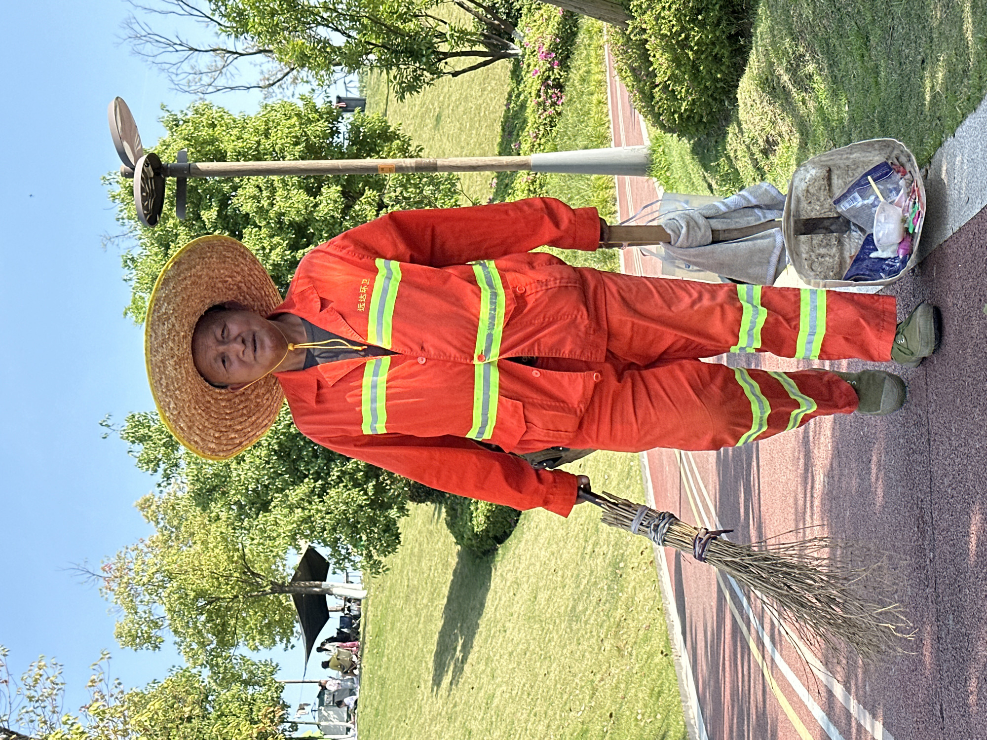 A man in orange reflective work clothes and a straw hat is standing outside in Jiangjin, China. He is holding a broom in his left hand, and his right hand is holding a shovel attached to a bin, which contains items of trash, over his shoulder. He is standing on a red running track next to a grassy field, with a group of people under a tent in the background. The sky is clear and blue.