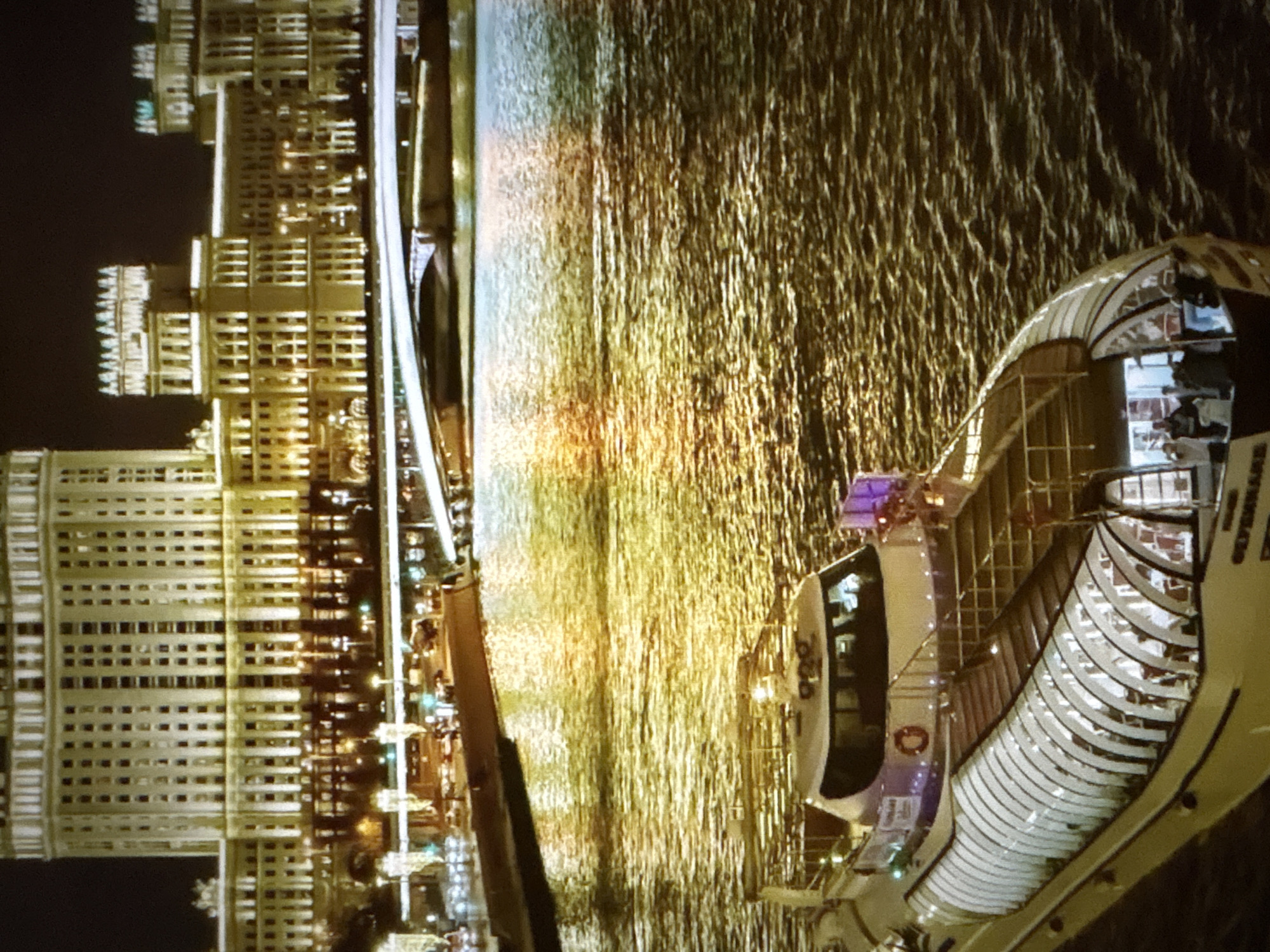 An aerial shot captures a nighttime scene of a river cruise boat with a glass enclosure sailing on dark, rippling water reflecting golden lights from a city skyline. The boat, partially visible, shows people inside its illuminated interior and a few standing on its upper deck. In the background, a large, ornate building with numerous windows and bright lights dominates the scene, suggesting an urban setting. A bridge spans across the water in front of the building. The image composition and lighting indicate a picturesque, albeit slightly blurry, view of a city at night, likely a popular tourist area. This scene does not depict the city of Pori, Finland.
