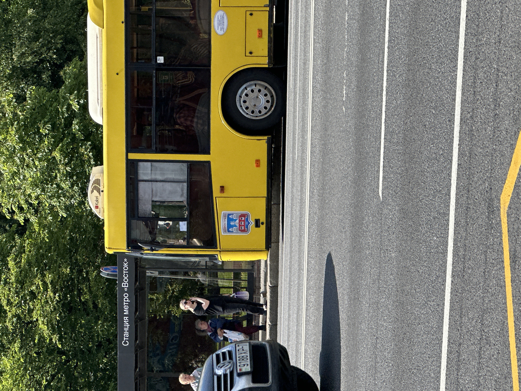 The image shows a yellow bus at a bus stop on a sunny day. The bus stop sign reads "Станция метро «Восток»" which translates to "Vostok metro station." Three people are visible at the bus stop. One woman, wearing a black top and black pants, is standing on her phone. Next to her, an older woman in a blue jacket carries a white bag. To the left of them, a person with short grey hair is partially visible. A silver car with a license plate reading "9188 XP-7" is parked to the left of the bus stop. The bus itself has a crest on its side with a city skyline and the word "МИНСКТРАНС" which means "Minsktrans." The background is filled with lush green trees, suggesting a location in a city park or along a tree-lined avenue. The road in the foreground has white and yellow lane markings.