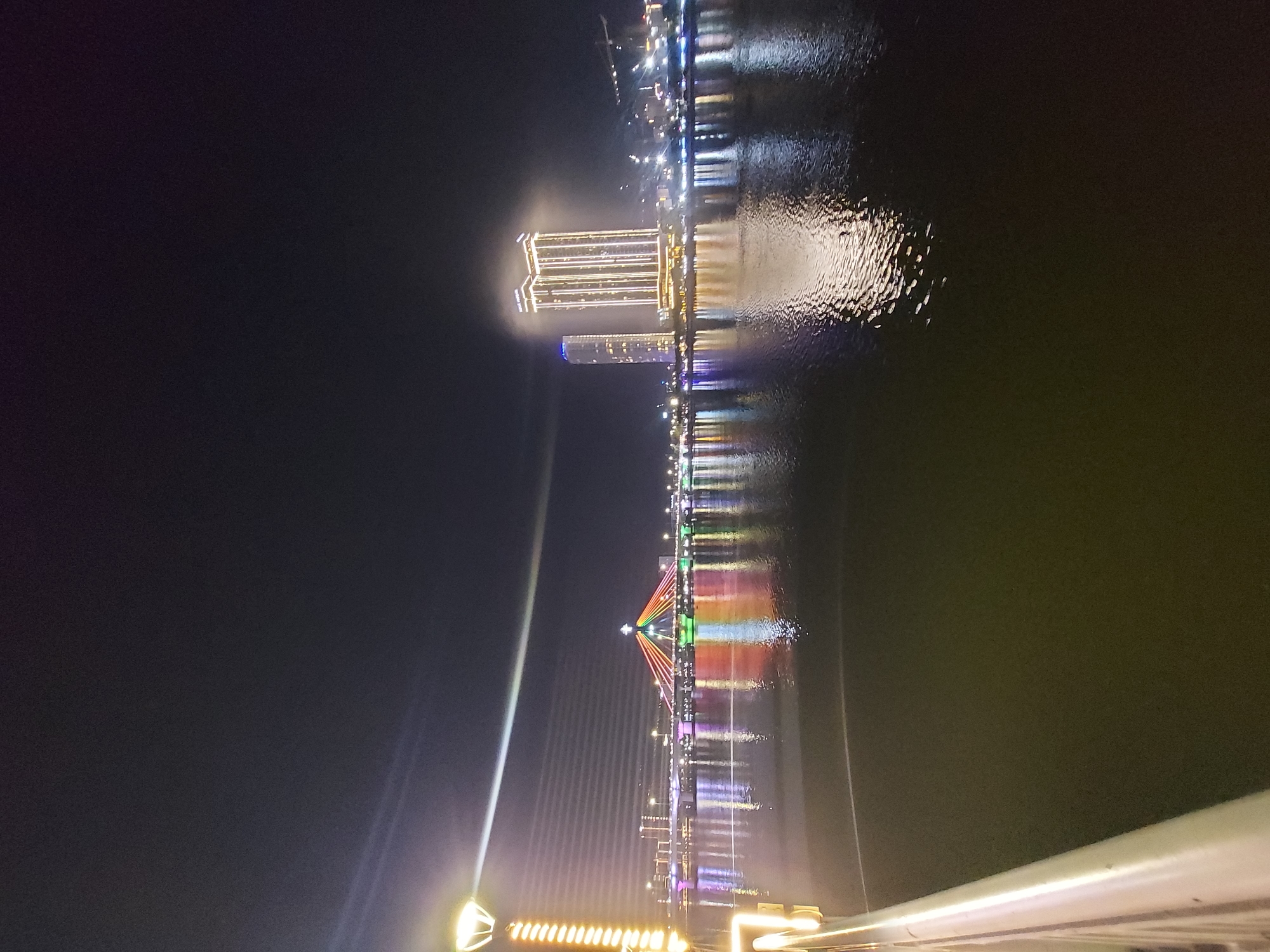 The image captures a nighttime view of the Han River in Đà Nẵng, Vietnam. The Dragon Bridge is illuminated with colorful lights, spanning across the river. On the right side of the image, two modern high-rise buildings, likely hotels or office spaces, stand illuminated, reflecting on the water's surface. A pedestrian bridge or walkway with a railing is visible in the foreground on the left, with a decorative lamp post. The dark sky is punctuated by the bright lights of the city and a beam of light emanating from the Dragon Bridge. The water of the Han River is dark and reflects the city lights.