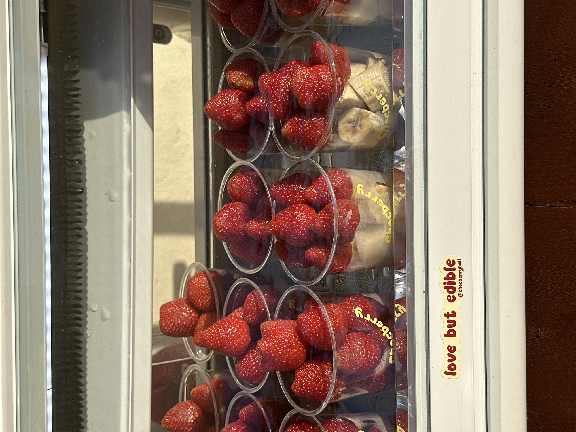 The image shows a close-up, vertical view of a refrigerated display case filled with clear plastic cups of strawberries and slices of banana. The display is lit from within, and the strawberries are bright red and appear fresh. Some of the cups are filled to the brim, while others have fewer fruits. Behind the fruits, yellow packaging with red text is visible, indicating the brand or product. One sticker on the exterior of the display case reads "love but edible" in red script with a yellow background. The setting appears to be a commercial establishment, possibly a food stall or shop, given the refrigerated display. The lighting suggests it could be daytime. The image is taken in North Kuta, Indonesia.