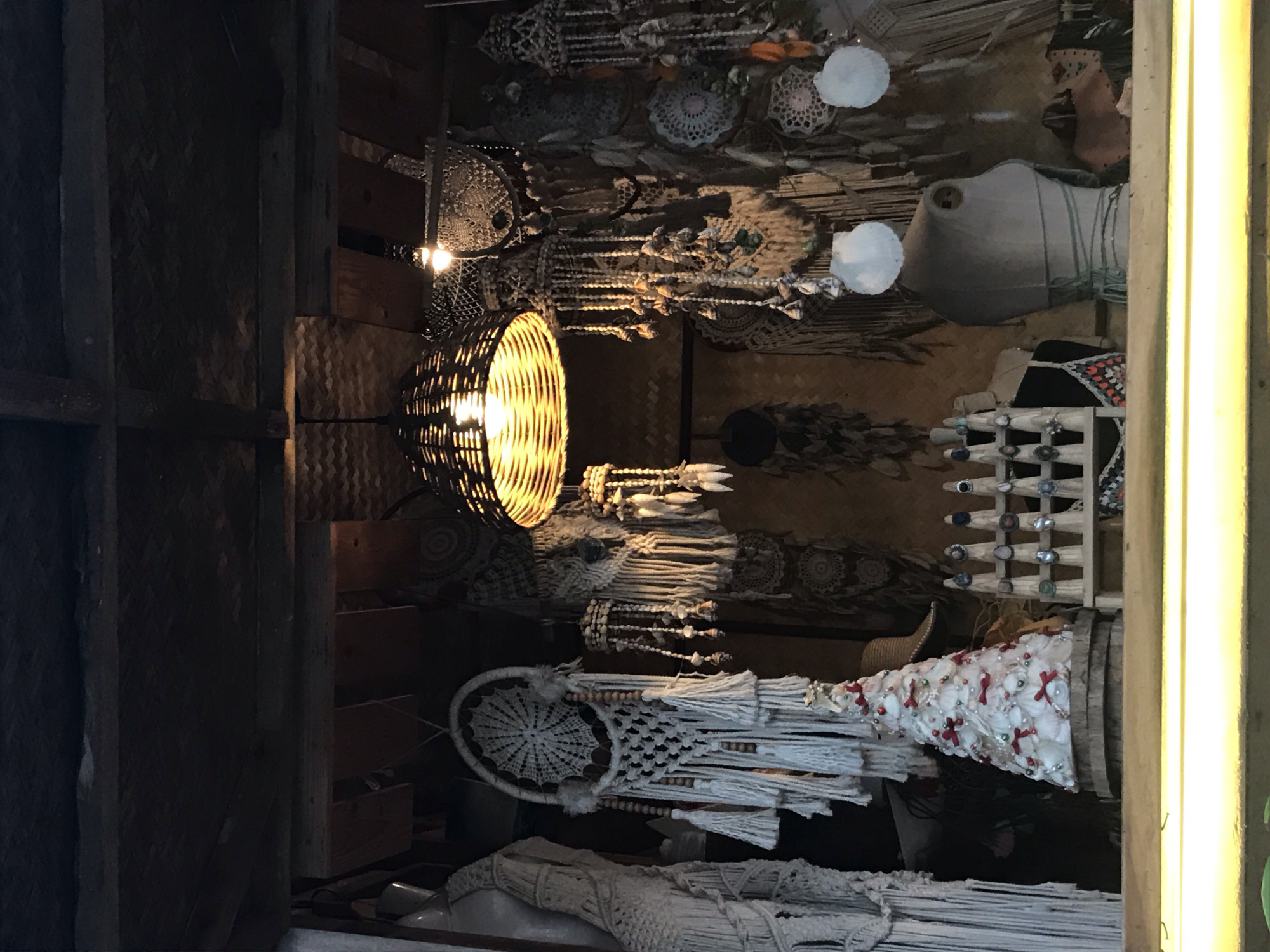 The image depicts a shop in Mũi Né, Vietnam, showcasing handcrafted decorative items. A wicker basket lamp is illuminated, alongside macrame dream catchers, seashell wind chimes, and ornaments. A cone-shaped object, adorned with shells and red bows, sits near a display of rings. The scene is partially framed by dark wooden structures and a bright vertical light fixture.