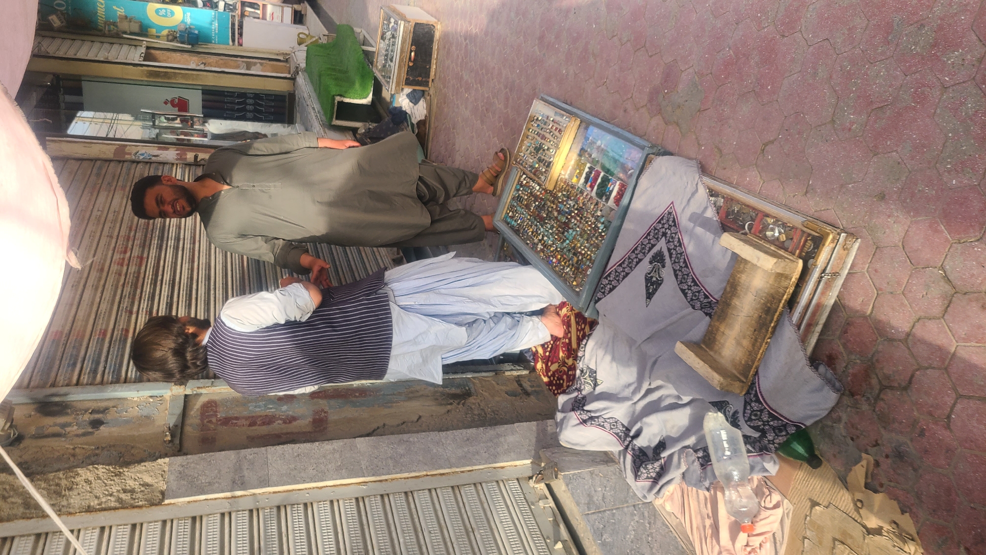 Two men are standing on a sidewalk in Quetta City Tehsil, Pakistan, next to a street market. The man on the left is wearing a blue and white striped vest over a white shirt and light blue shalwar. He has long, dark hair. The man on the right is wearing a dark green shalwar kameez. He has short, dark hair and a beard. They appear to be in conversation. In front of the man on the left, there is a display of rings and other jewelry laid out in trays on the ground. A light-colored blanket with a dark pattern is spread on the ground next to the jewelry. A wooden object, possibly a stand, rests on top of the blanket and some of the jewelry trays. Behind the men, there is a closed storefront with a corrugated metal shutter. To the right of the storefront, a green fabric-covered object is visible, along with a wooden box and more display trays containing jewelry. A plastic water bottle and a green bottle are lying on the blanket. The ground is paved with red hexagonal tiles.
