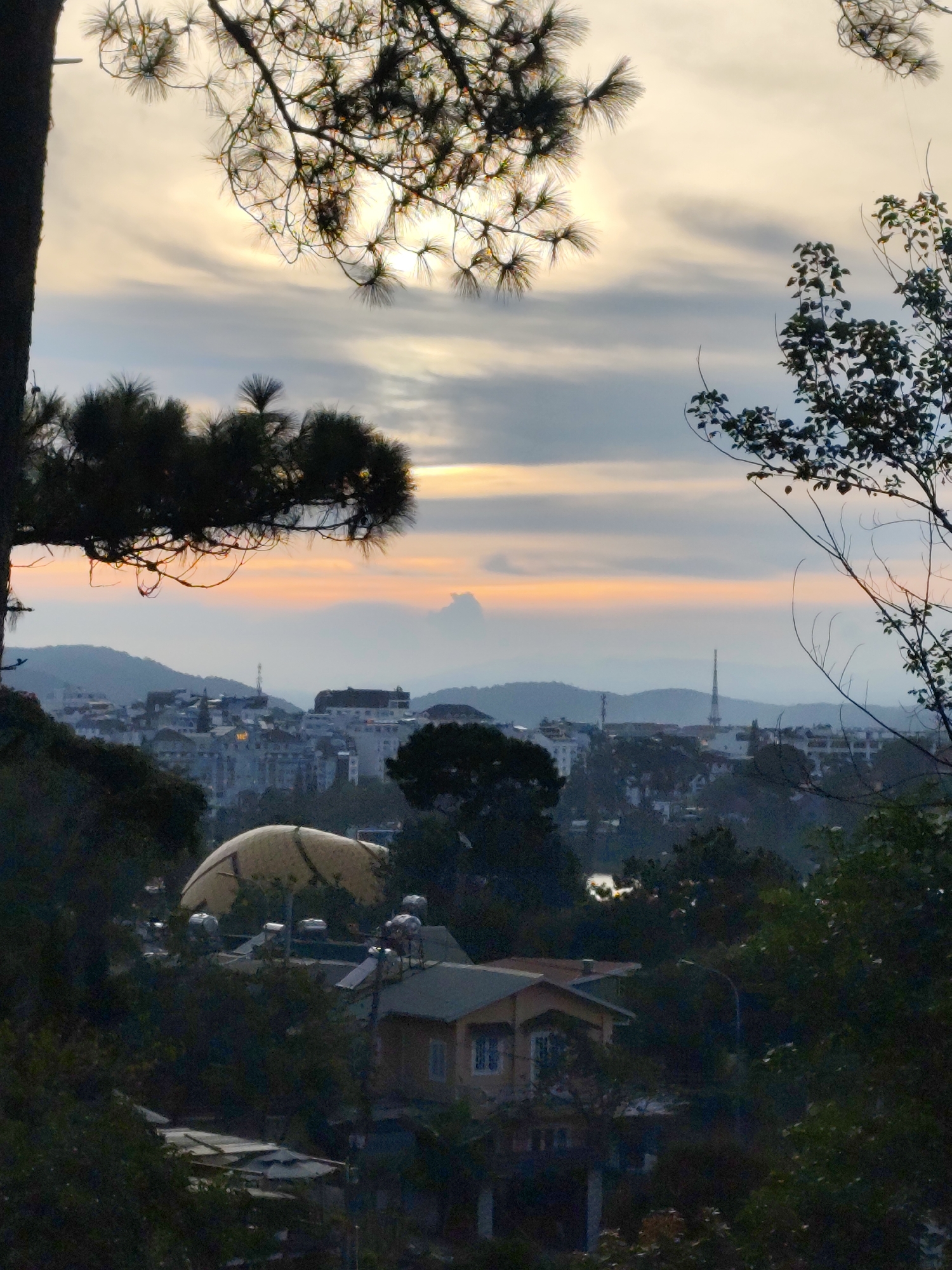 The image captures a serene view of Da Lat, Vietnam, during what appears to be either sunrise or sunset. In the foreground, lush green foliage and the dark silhouette of pine tree branches frame the scene. The sky is a blend of soft grays, whites, and a warm orange hue near the horizon, suggesting a twilight atmosphere.
Further into the frame, a city unfolds with buildings of varying sizes, nestled among rolling hills. The architecture suggests a European influence, with some structures displaying white facades and tiled roofs. A prominent feature is a large, light-colored domed structure, possibly a public building or landmark. Several solar water heaters are visible on the rooftops of some houses, indicating a common feature of local dwellings.
In the middle ground, the city's skyline is dotted with taller buildings and a distinctive telecommunications tower. The overall impression is one of a peaceful urban landscape gradually emerging from or receding into the soft light of dawn or dusk. The atmosphere is tranquil, with no visible human activity, emphasizing the quiet beauty of the natural and built environment.