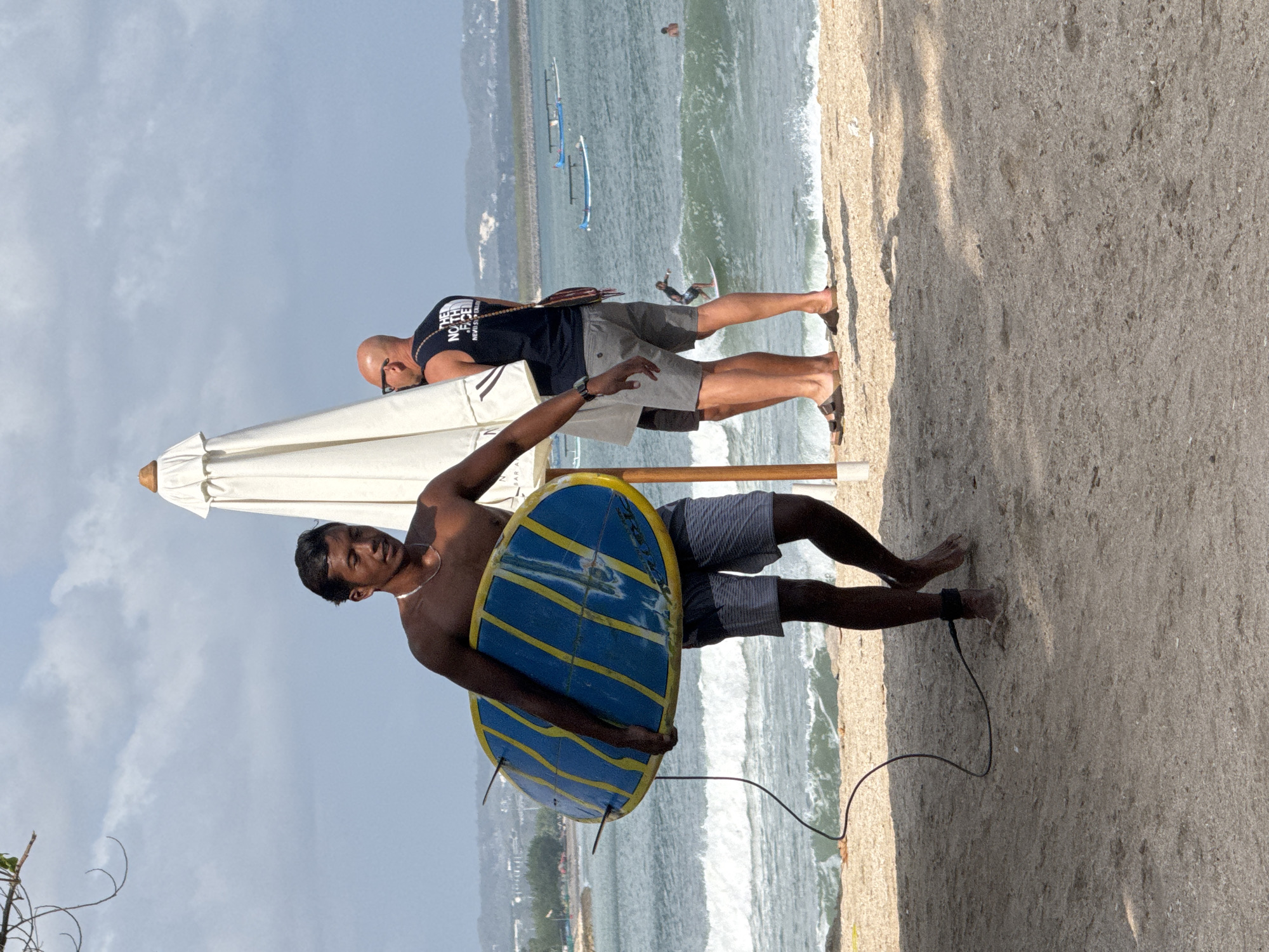 A young man, shirtless and wearing striped shorts, walks across a sandy beach in Kuta, Indonesia, carrying a blue and yellow striped surfboard. A coiled black leash hangs from the board. Behind him, two men stand near a large white beach umbrella. One man, wearing sunglasses and a black t-shirt, looks out at the ocean. The other man, further back, is partially obscured. In the background, the ocean has moderate waves, and several small boats are visible. The sky is partly cloudy.