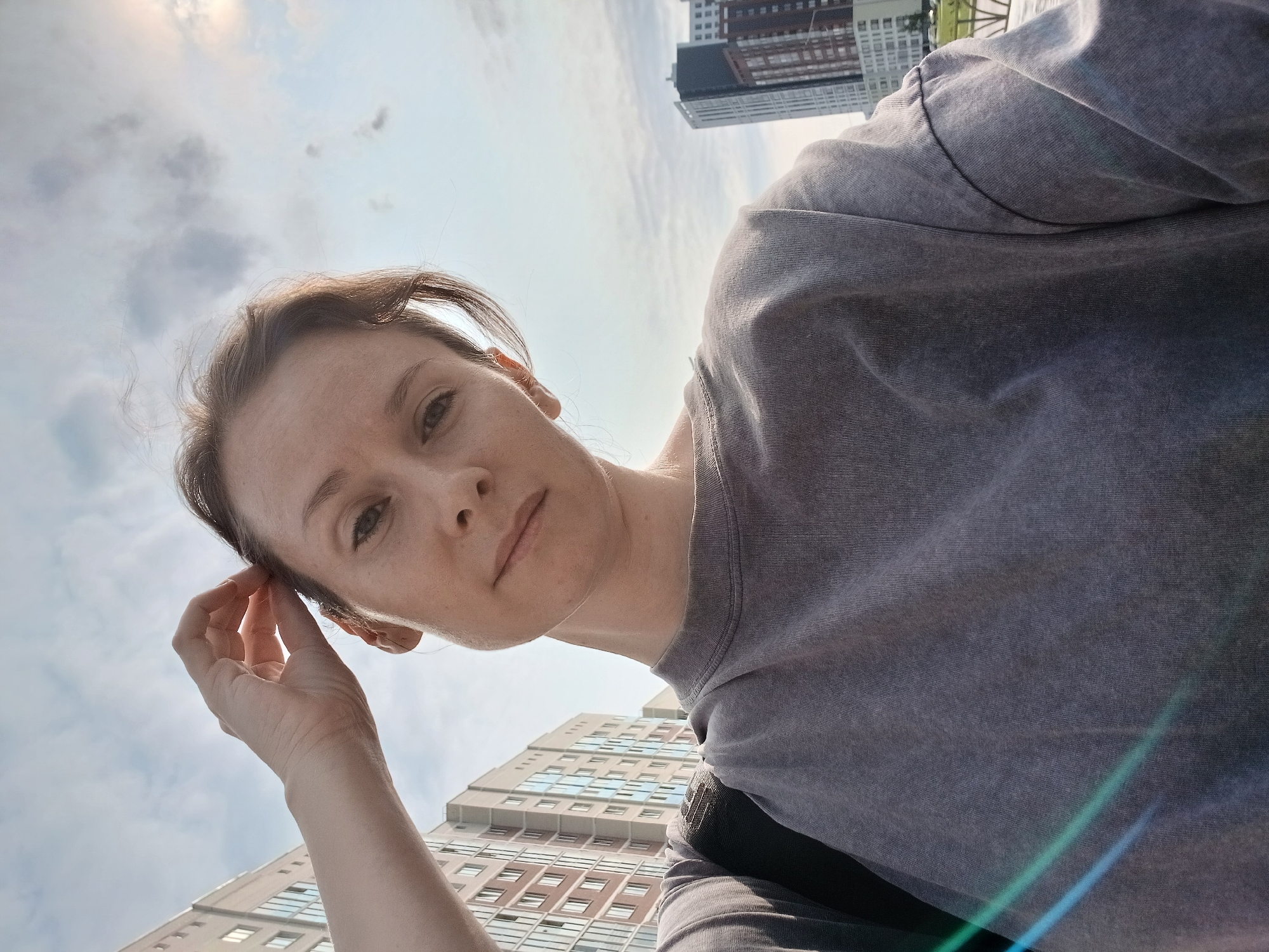 A low-angle, close-up selfie of a young Caucasian woman with light brown hair and fair skin is taken outdoors in Yekaterinburg, Russia. She is looking directly at the camera with a neutral expression, and her left hand is raised to her head, with her fingers touching her hair. She is wearing a gray, oversized sweatshirt, and a black backpack strap is visible over her left shoulder.
The background features a bright, cloudy sky, and two multi-story apartment buildings. The building on the left is pale brown with a repeating pattern of windows. The building on the right is taller and features a dark top section and brick-patterned lower floors. Some light flares or lens flares are visible on the left side of the image, suggesting it was taken during daylight hours. The overall impression is an urban environment, possibly a residential area.