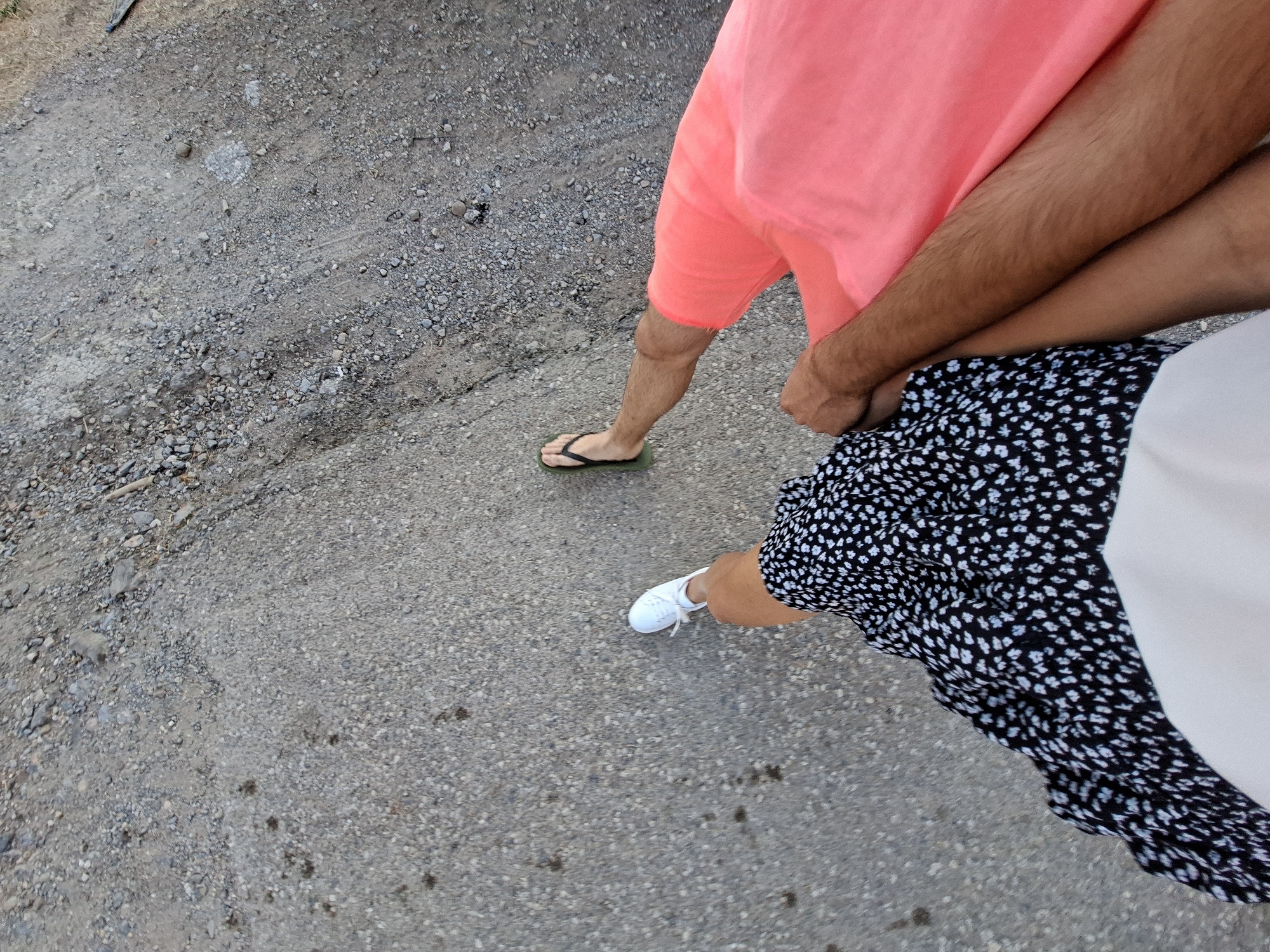 An overhead shot shows two people holding hands and walking on a gravel path. The person on the left wears a black and white floral dress and white sneakers. The person on the right wears a coral-colored pair of shorts and green flip-flops. The path is made of small gray pebbles and dirt. The scene is outdoors and appears to be in a rural or undeveloped area.